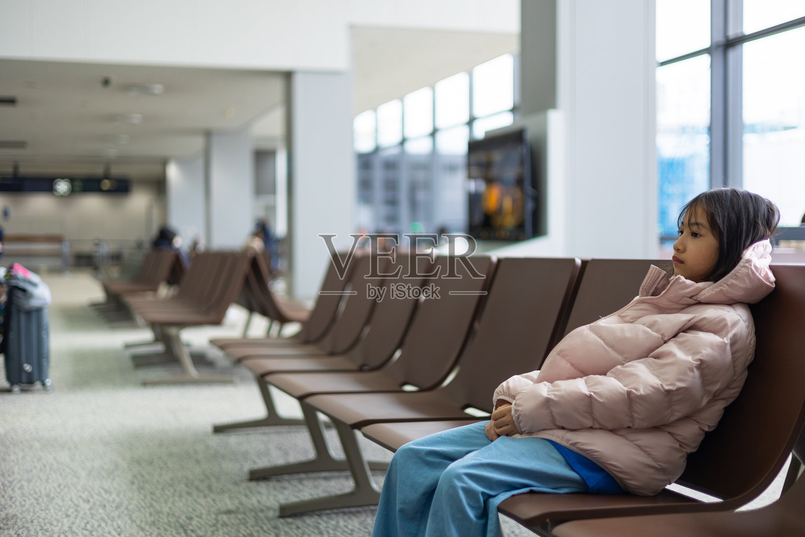 穿着冬季外套的可爱亚洲小女孩独自坐在机场休息室，沉浸在思考中。静谧的环境、空荡的椅子和柔和的灯光展现了孤独、耐心和未来的旅程。照片摄影图片