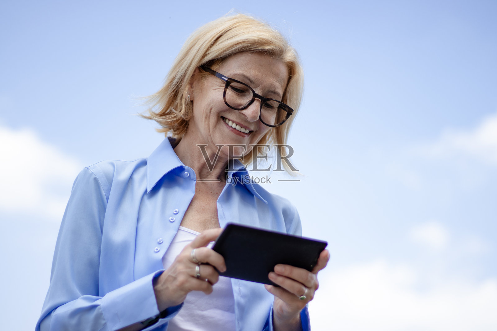 成熟的金发快乐女商人戴着眼镜，在建筑物的露台上休息，使用平板电脑。照片摄影图片