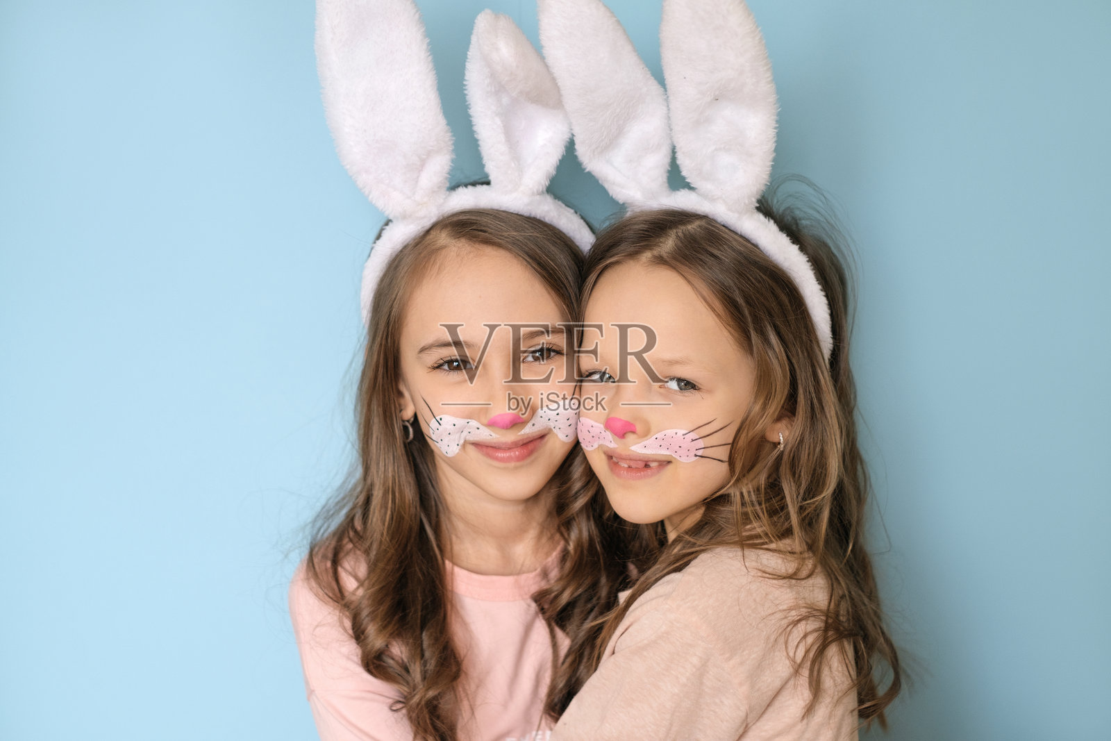 可爱的女孩们脸上画着兔子脸，头上戴着兔耳朵，互相拥抱，甜美地看着镜头。复活节假期。照片摄影图片