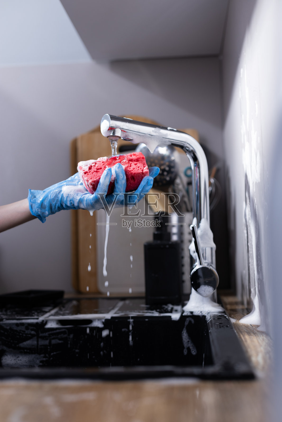 现代厨房水槽中，手握着一块沾满肥皂的粉色海绵，水流淌而下，黑色时尚的台面和干净的环境映衬着这一幕。照片摄影图片