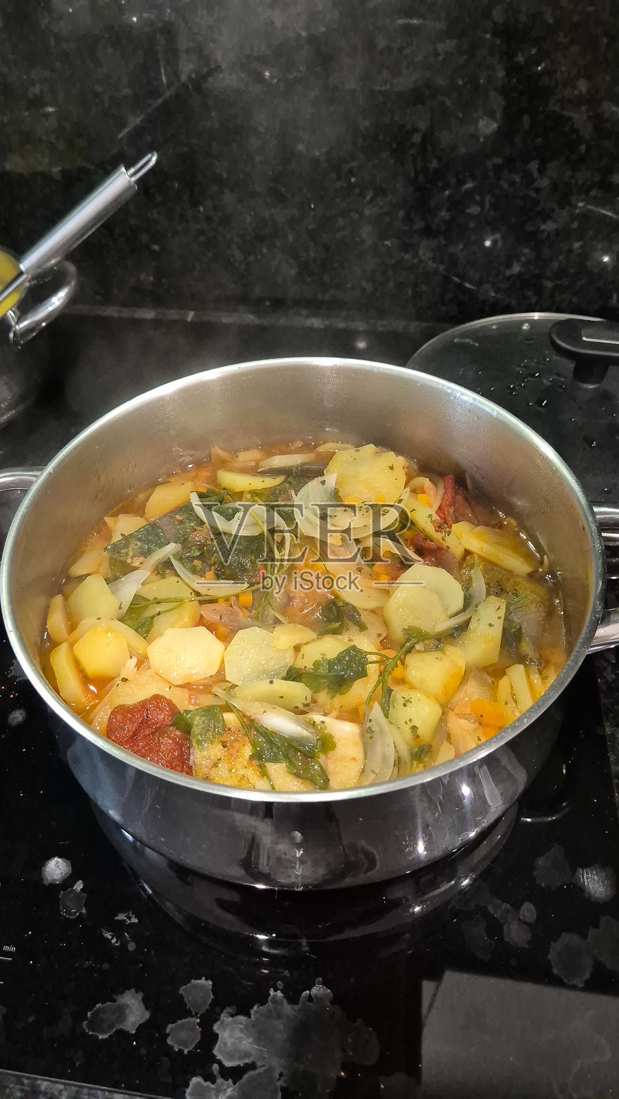 传统葡萄牙鳕鱼炖菜，称为bacalhau caldeirada，使用不锈钢锅烹制，配有切片土豆、洋葱、西红柿、辣椒、香草和橄榄油。在炉灶上制作的家常菜。照片摄影图片