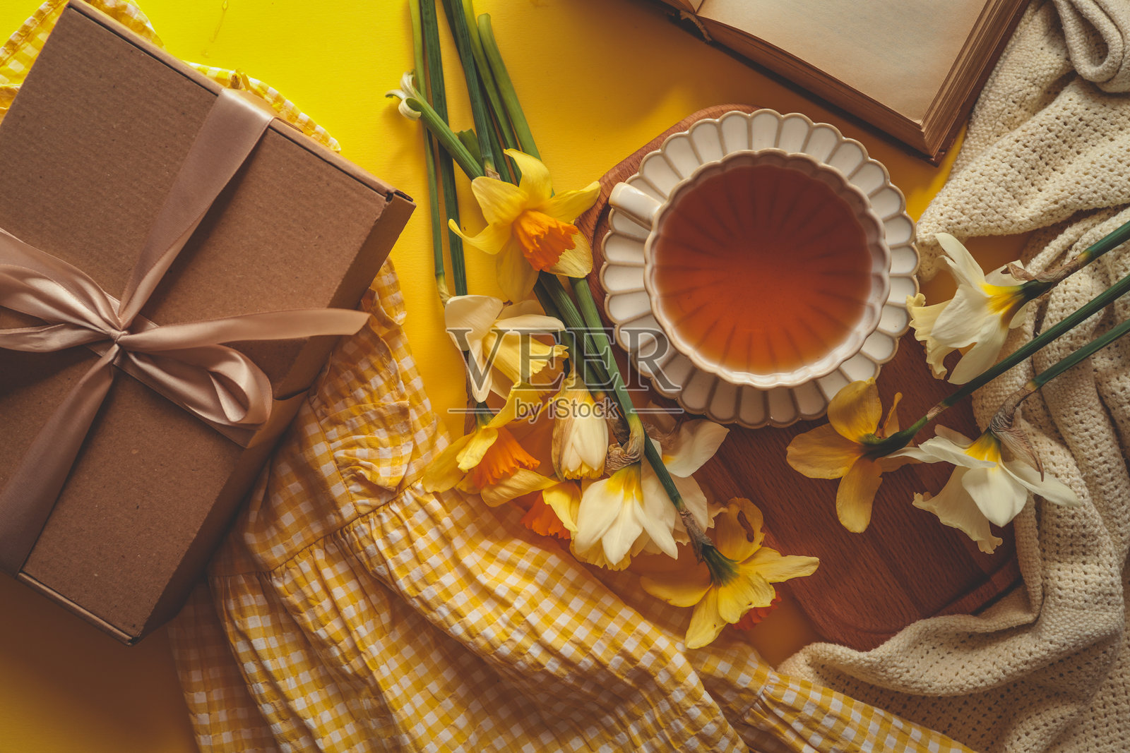 舒适春季场景的平铺图，背景为黄色，包含一杯茶、黄水仙花、一个带丝带的礼品盒和一本打开的书，周围有柔软的面料和格子布。照片摄影图片
