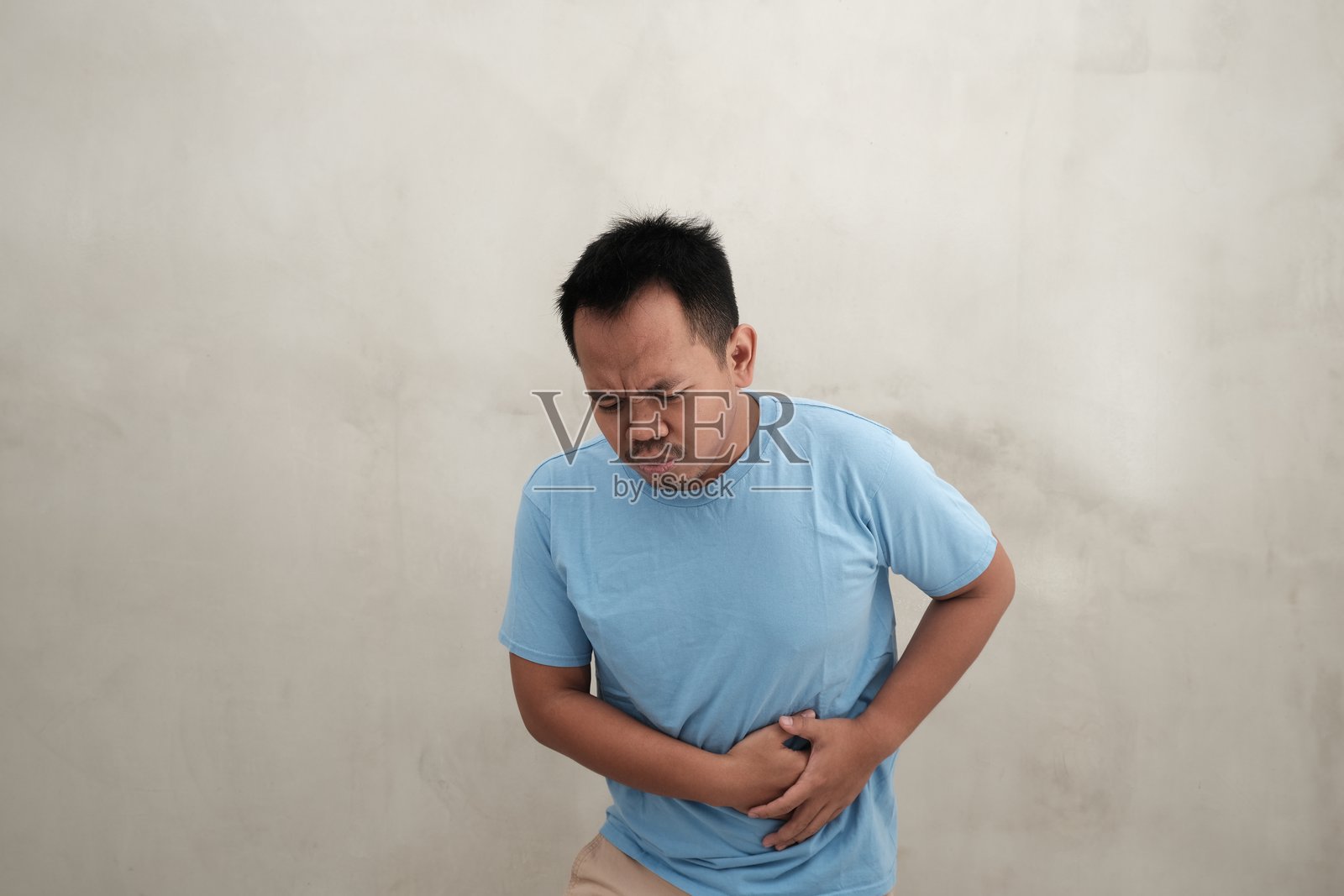 一位东南亚男子穿着浅蓝色T恤，明显感到不适，双手捂着肚子，正在忍受腹痛。照片摄影图片