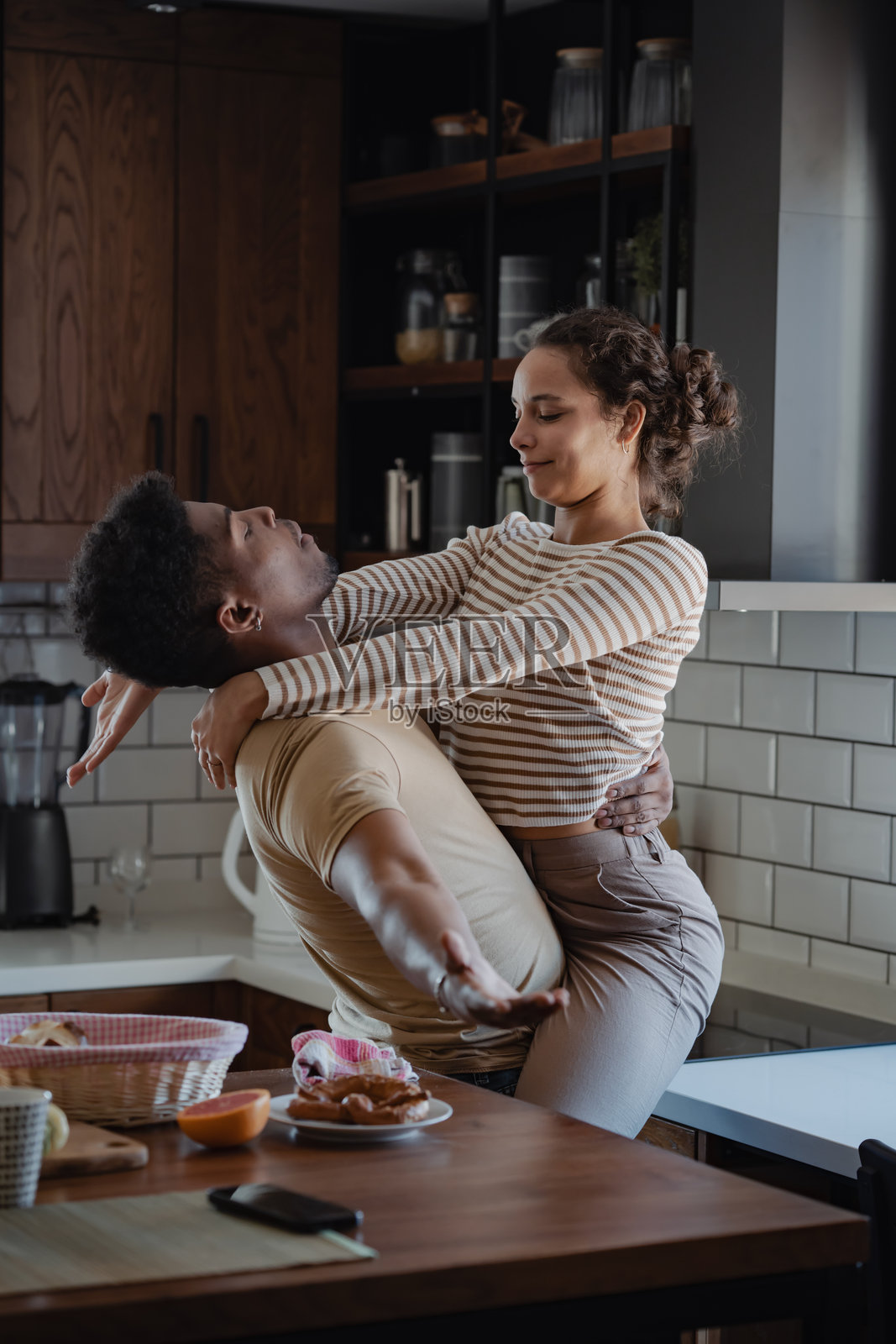 一对恩爱情侣在温馨的厨房中分享亲密时刻，男人将女人抱起，两人温暖地相视而笑。木质台面上可见早餐的摆设。照片摄影图片