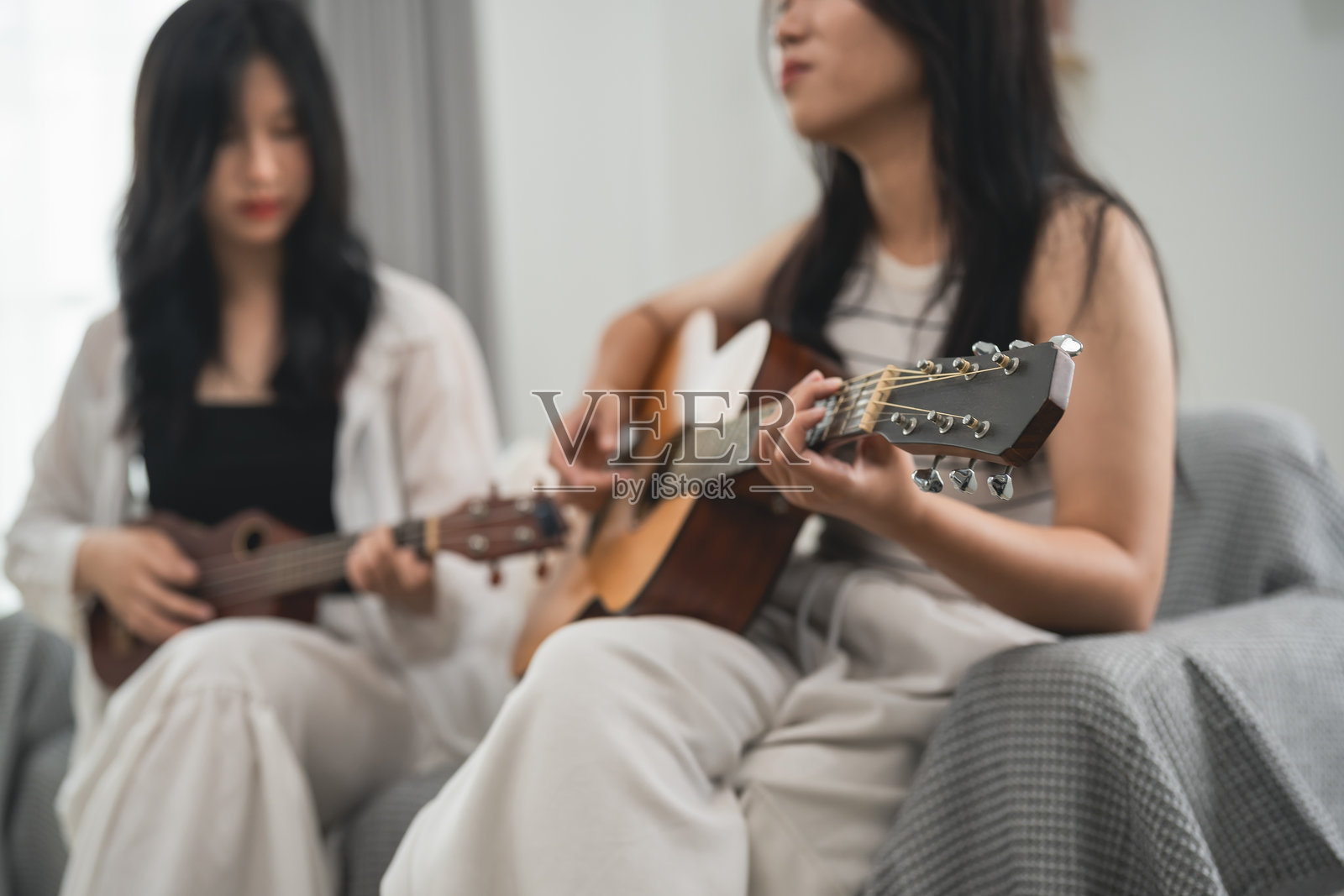 两位亚洲女性在温馨的室内环境中一起弹吉他和尤克里里，表达快乐，创造音乐，享受自然光带来的温暖氛围。照片摄影图片