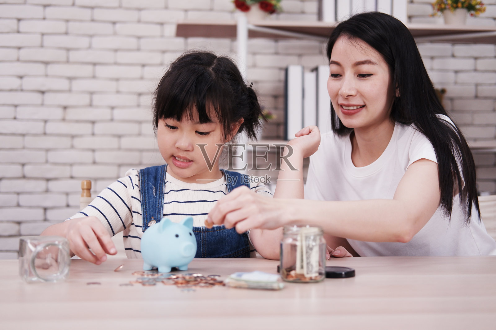 微笑的亚洲母女在图书馆的木桌上将硬币放入储蓄罐，为未来存钱。儿童教育，适合家庭教育的概念。照片摄影图片