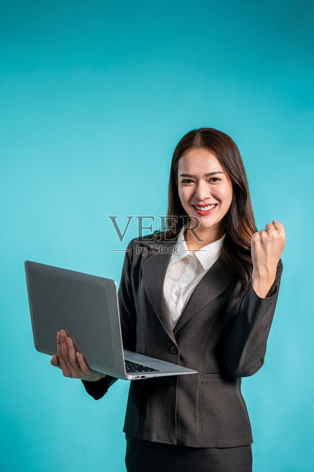 身穿正式服装的亚洲女商人在孤立的蓝色背景前站立，吸引人的年轻女性员工正在使用笔记本电脑并微笑，朝着镜头看，工作室屏幕截图。照片摄影图片