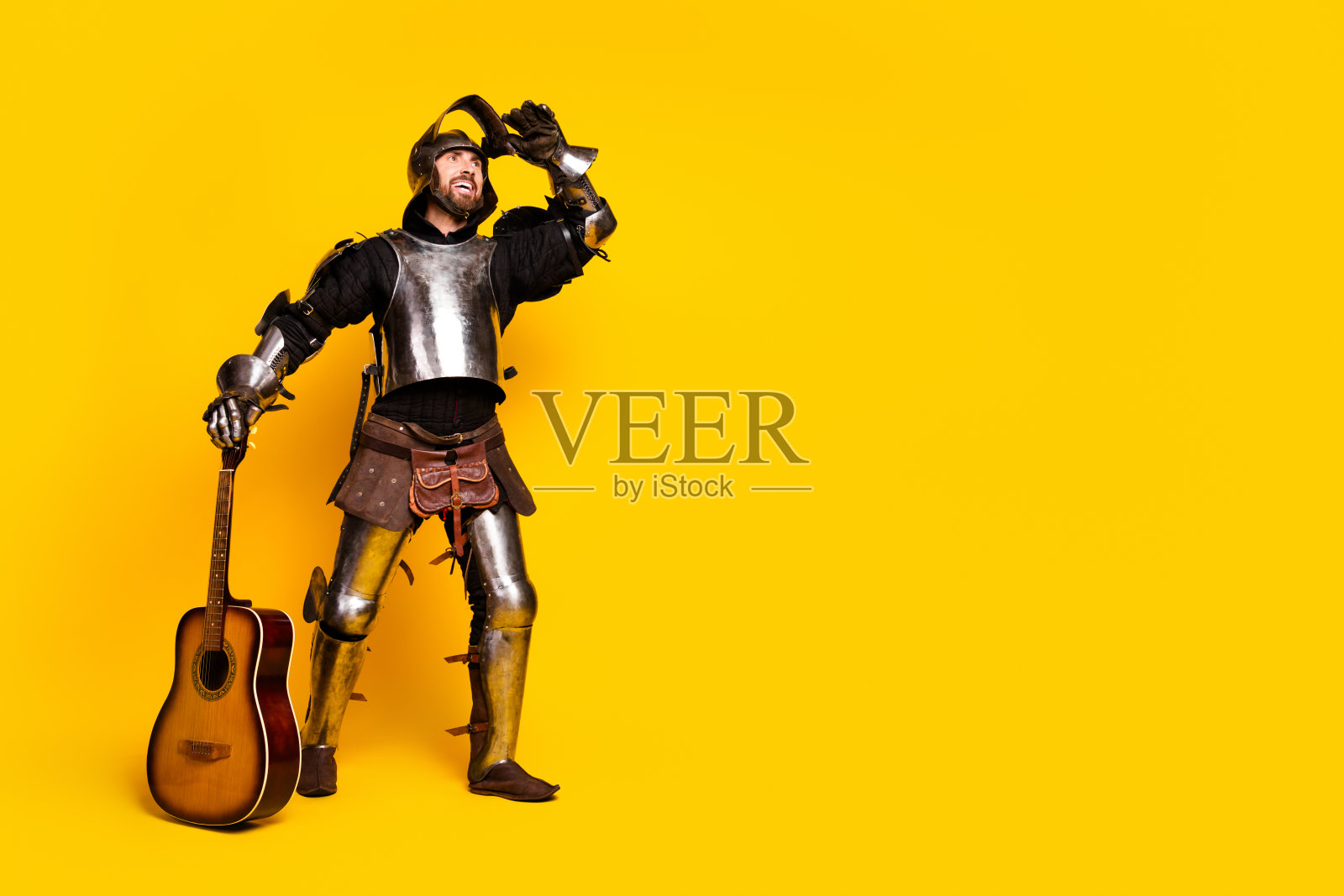 身穿中世纪骑士盔甲的年轻男子，手持吉他，背景为鲜艳的黄色。照片摄影图片