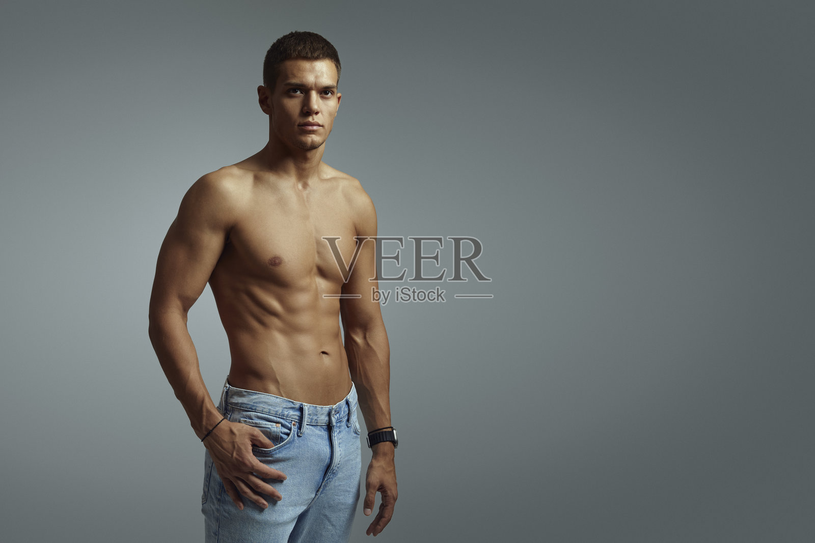 身材健美的年轻男子穿着牛仔裤，自信地摆姿势，背景为灰色，右侧留有空白空间。照片摄影图片