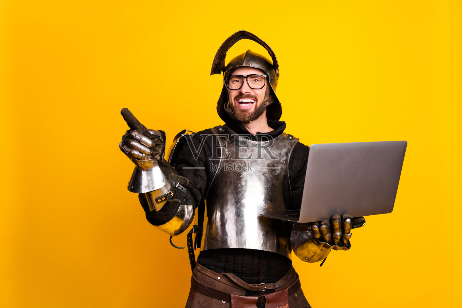 身穿骑士盔甲的年轻男子手持笔记本电脑，指向黄色背景。照片摄影图片