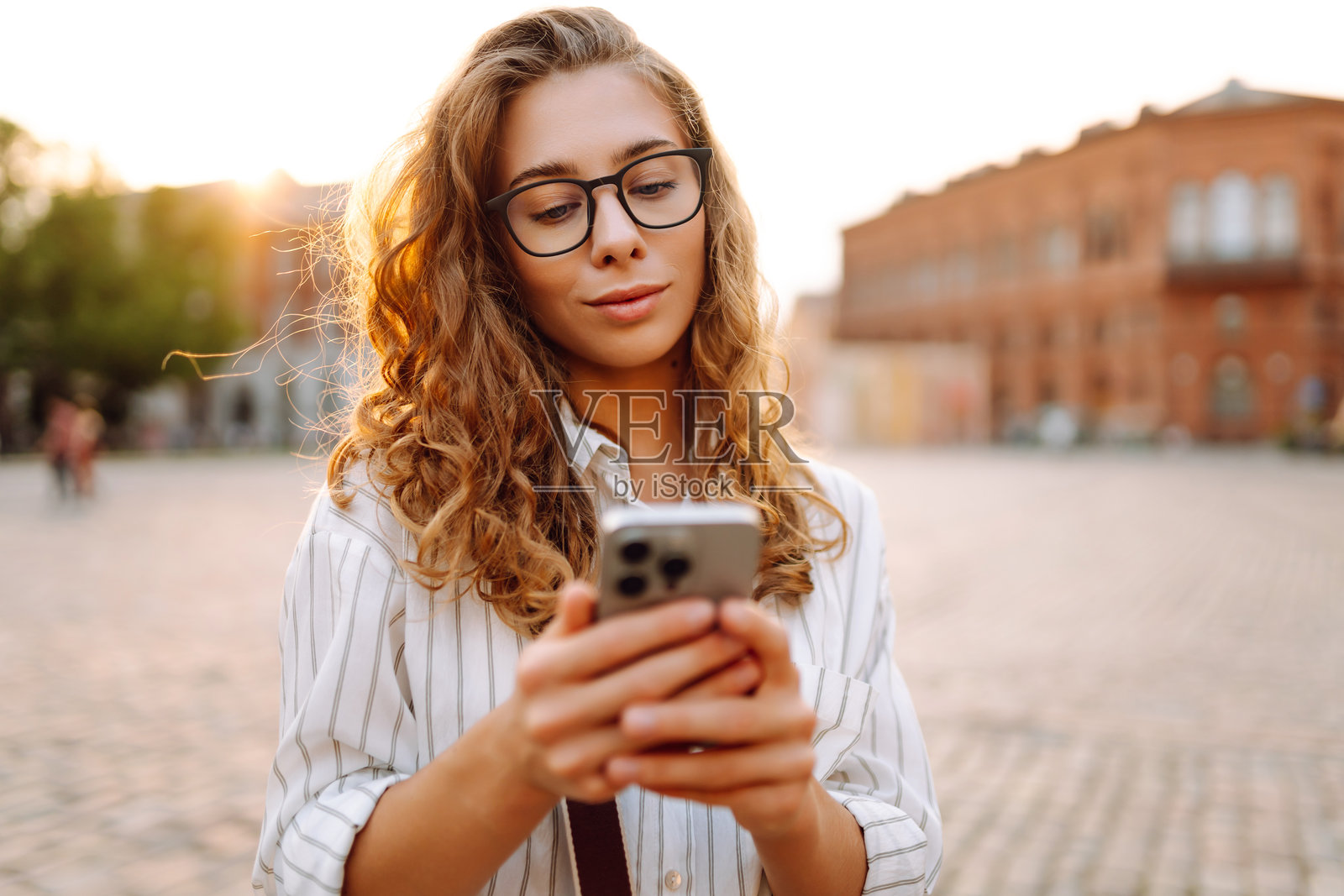 一位年轻女性穿着时尚的衣服，手握手机，走在阳光明媚的城市街道上。照片摄影图片