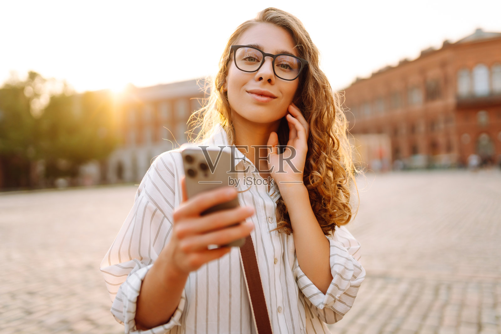 一位年轻女性穿着时尚的衣服，手握手机，走在阳光明媚的城市街道上。照片摄影图片