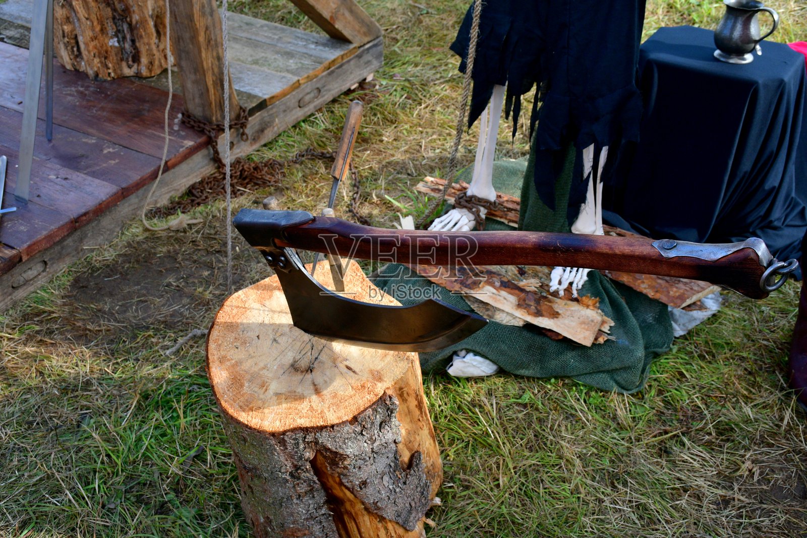 一把巨大的斧头近距离特写，木柄插入一根巨大的原木，旁边是一个骷髅的复制品和其他中世纪设备，拍摄于夏季的波兰。照片摄影图片