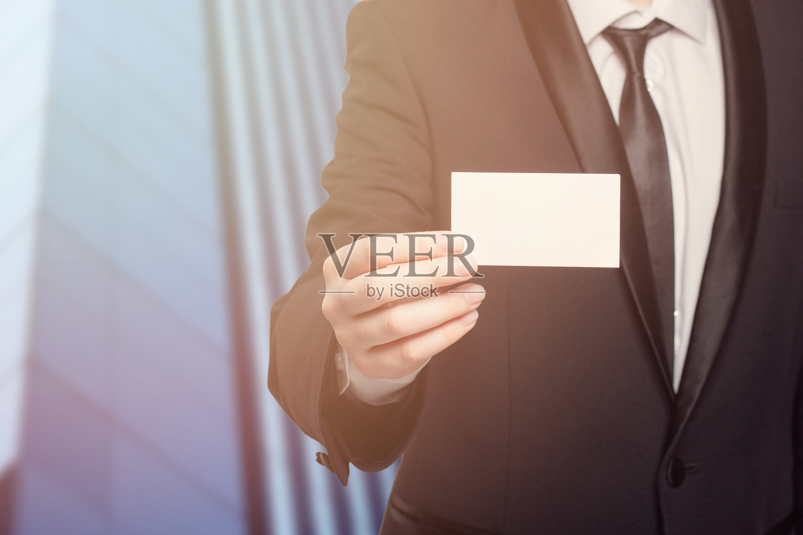 一位身穿西装的男士在玻璃摩天大楼背景下手持一张白色名片。照片摄影图片