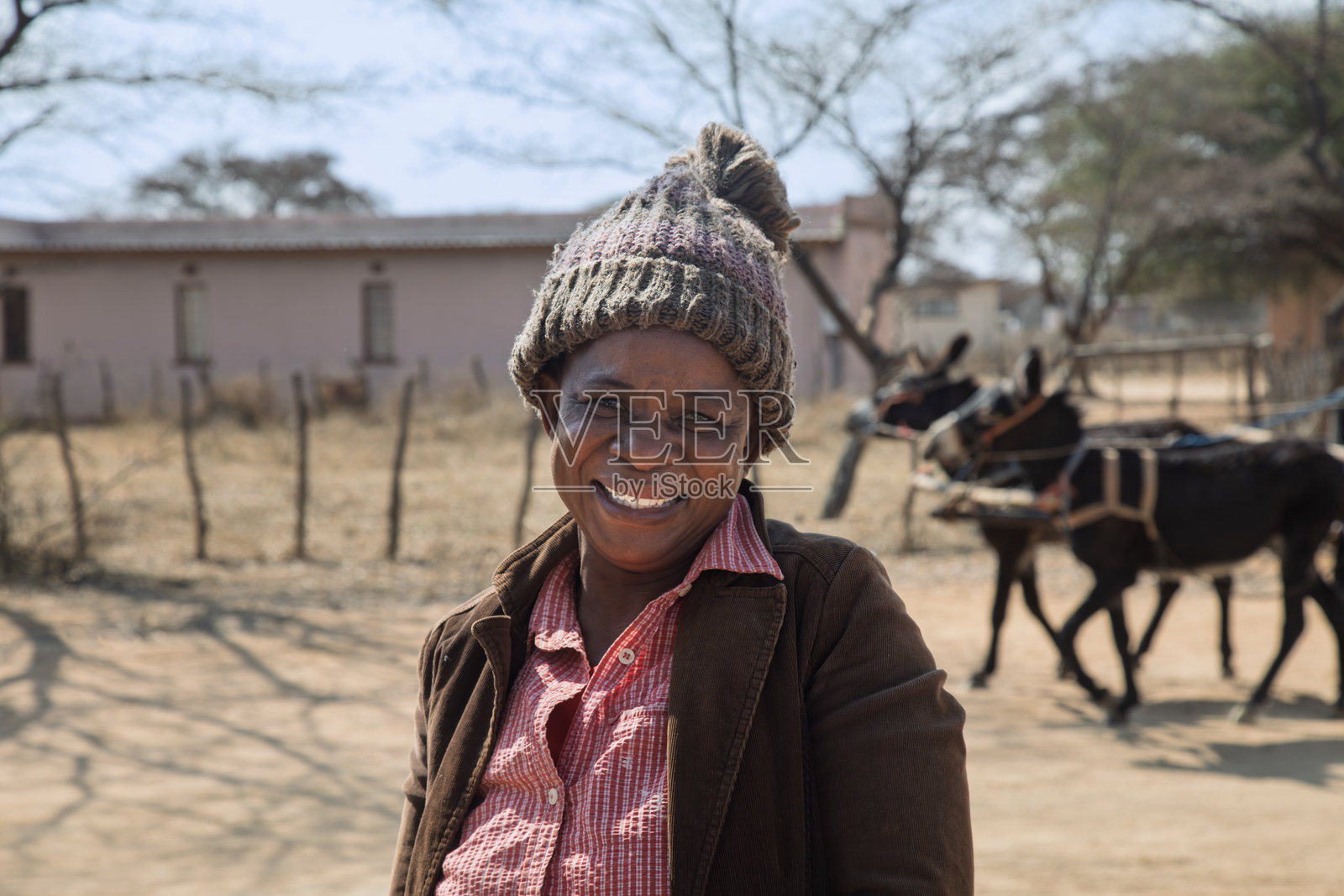 非洲村庄，快乐大笑的单身非洲女性肖像，戴着毛线帽，博茨瓦纳乡村，户外泥土路上有驴车经过照片摄影图片
