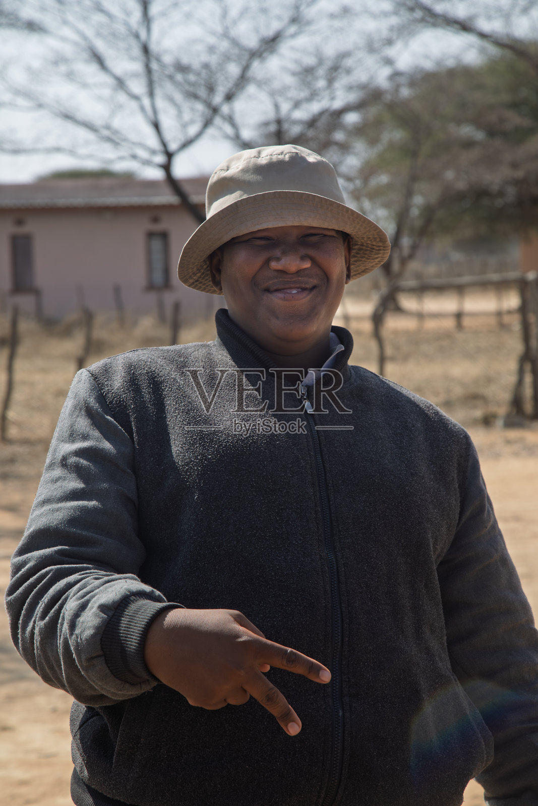 快乐的年轻非洲男性肖像，超重，做出胜利手势，博茨瓦纳乡村地区，户外庭院，背景有房屋。照片摄影图片