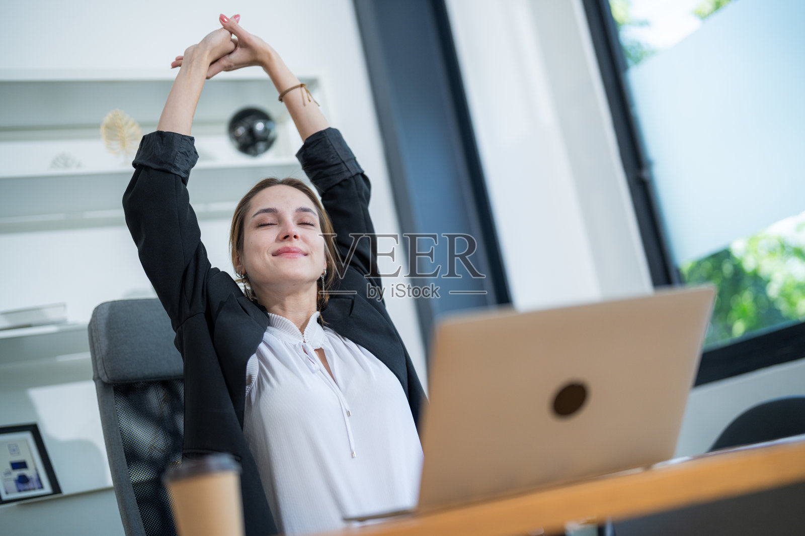 女性在明亮的办公室空间中休息时，在桌子前伸展身体，面前放着一台打开的笔记本电脑。照片摄影图片