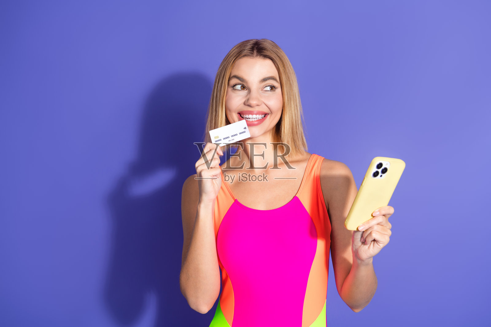 穿着明亮运动服的快乐女性，手持手机和信用卡，时尚地摆姿势，背景为鲜艳的紫色。照片摄影图片