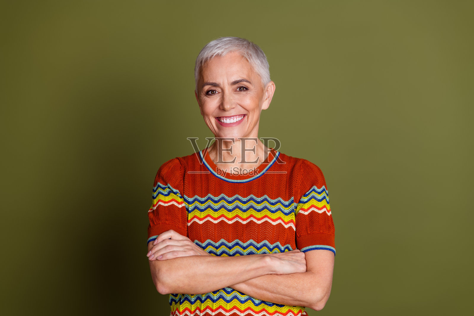 微笑的老年女性，短白发，穿着彩色条纹衬衫，站在卡其色背景前。照片摄影图片