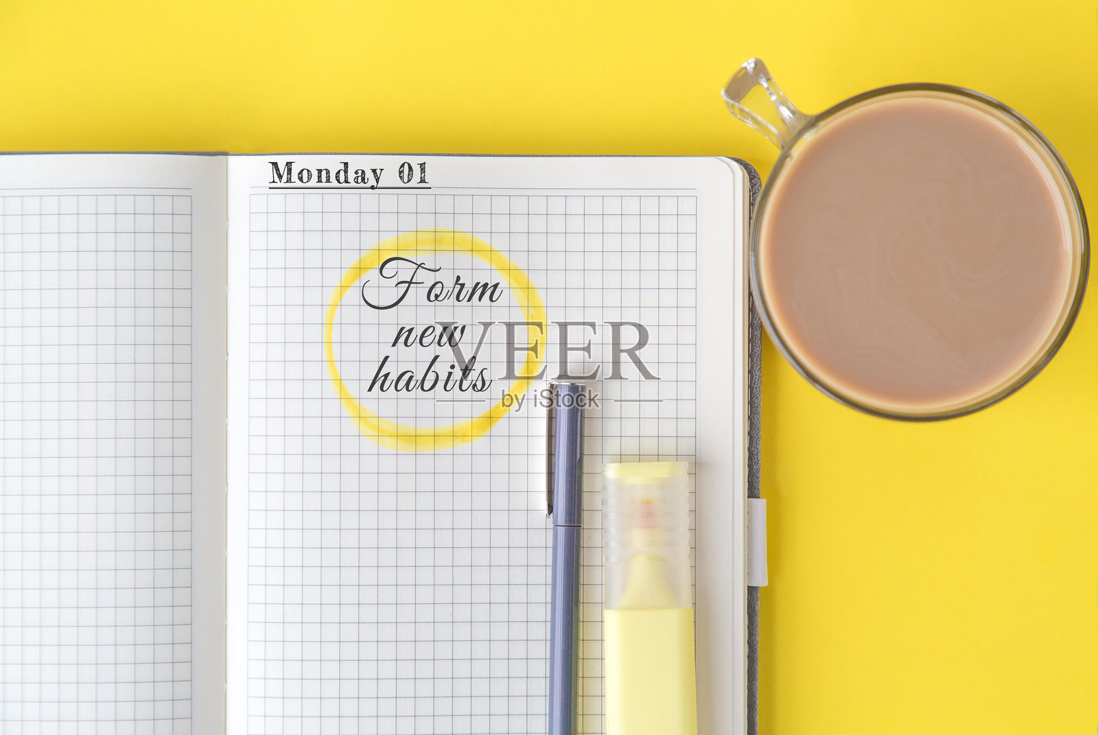 打开的格子纸笔记本，显示着用黄色圈出的激励短语“养成新习惯”，旁边有一支钢笔、一支荧光笔和一杯可可，背景为黄色，体现自我提升的概念。照片摄影图片