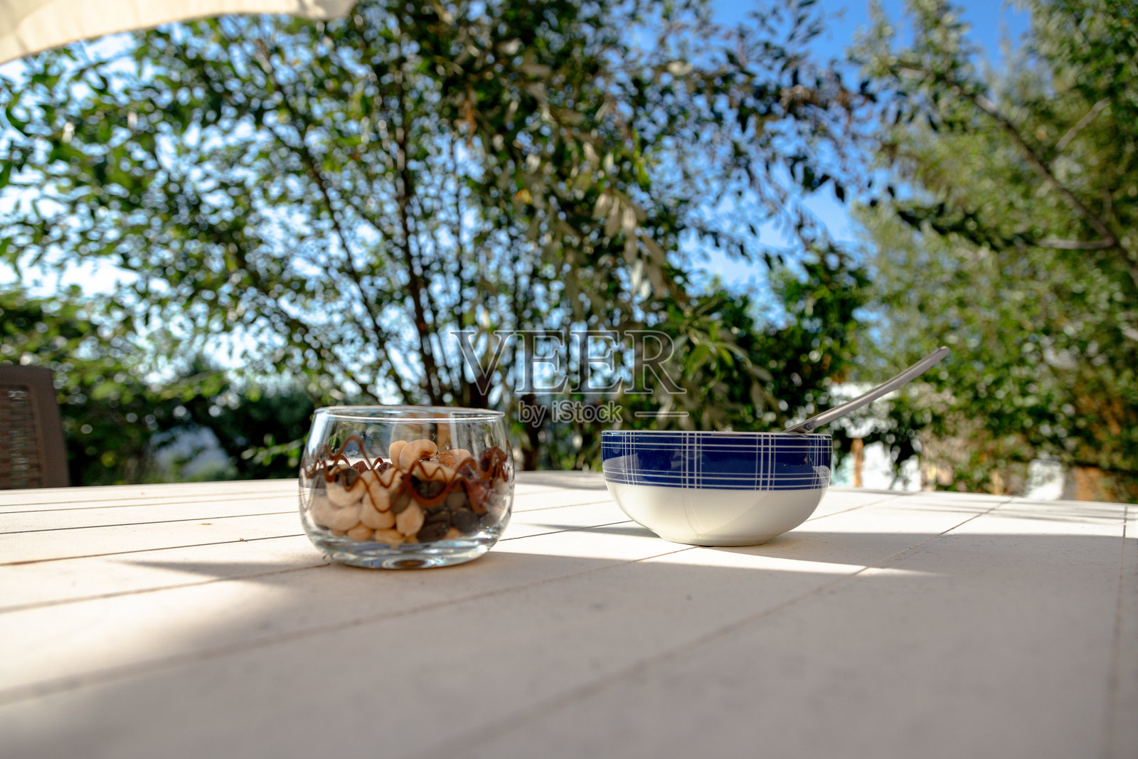 一个装满干果、坚果、腰果、榛子、南瓜子、葡萄干和杏仁的玻璃碗，放在户外桌子上，旁边有叠放的蓝白色碗。在明亮的阳光下享受轻松的零食时光。照片摄影图片