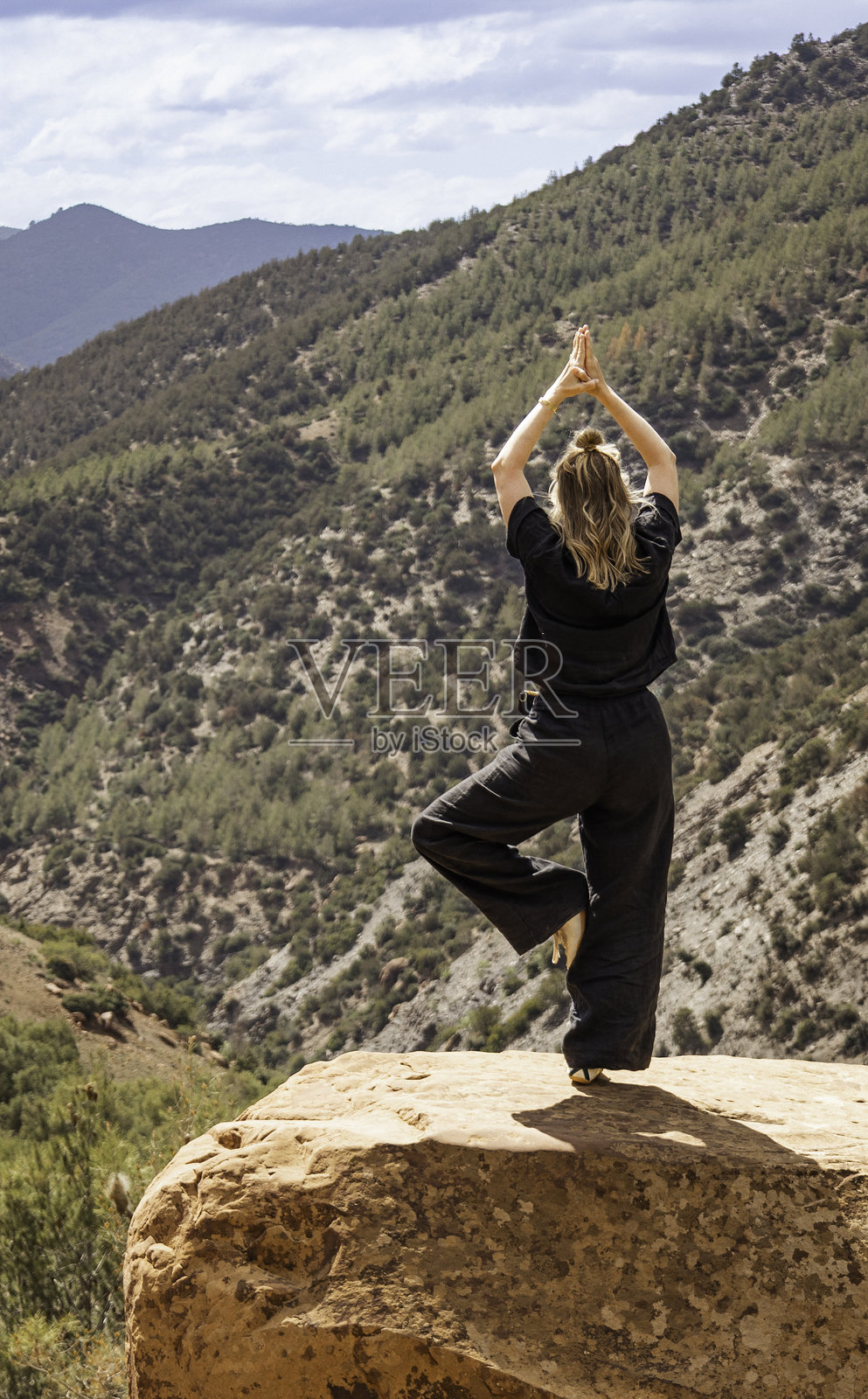 一位女性在摩洛哥阿特拉斯山脉的乌里卡谷，岩石上练习树式瑜伽，俯瞰森林覆盖的山脉。照片摄影图片