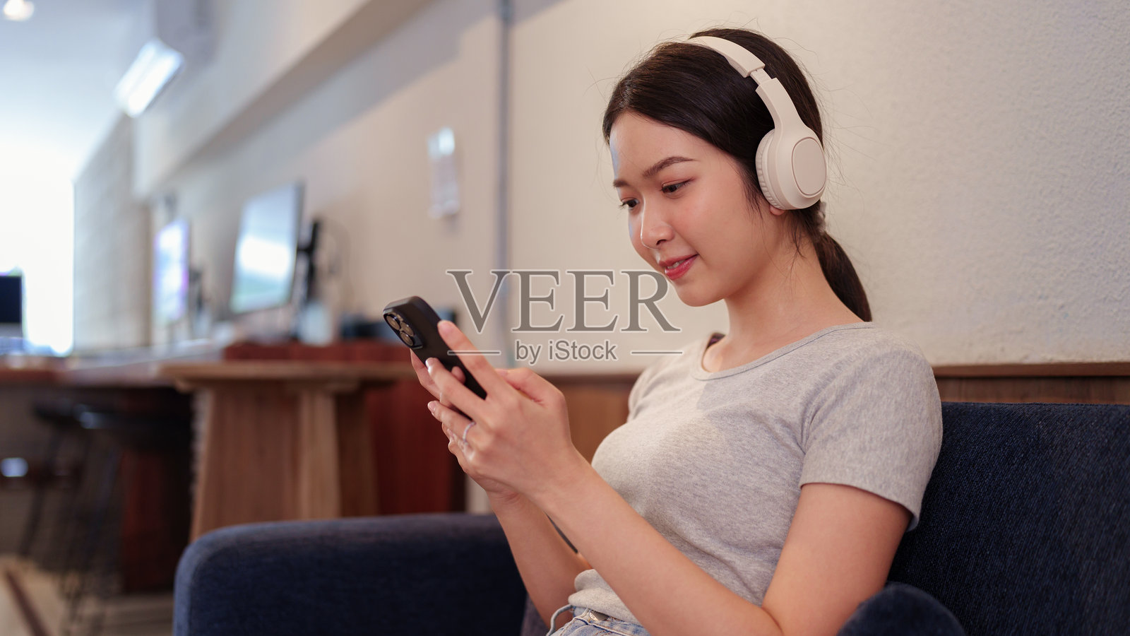 年轻女性坐在沙发上放松，戴着耳机听音乐，同时在智能手机上浏览歌曲。照片摄影图片