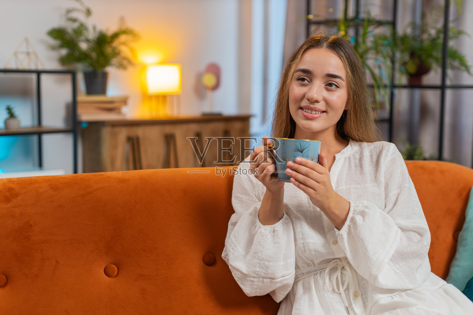 微笑的年轻女性坐在沙发上，早晨在家里喝着一杯温暖的咖啡或草本茶。照片摄影图片