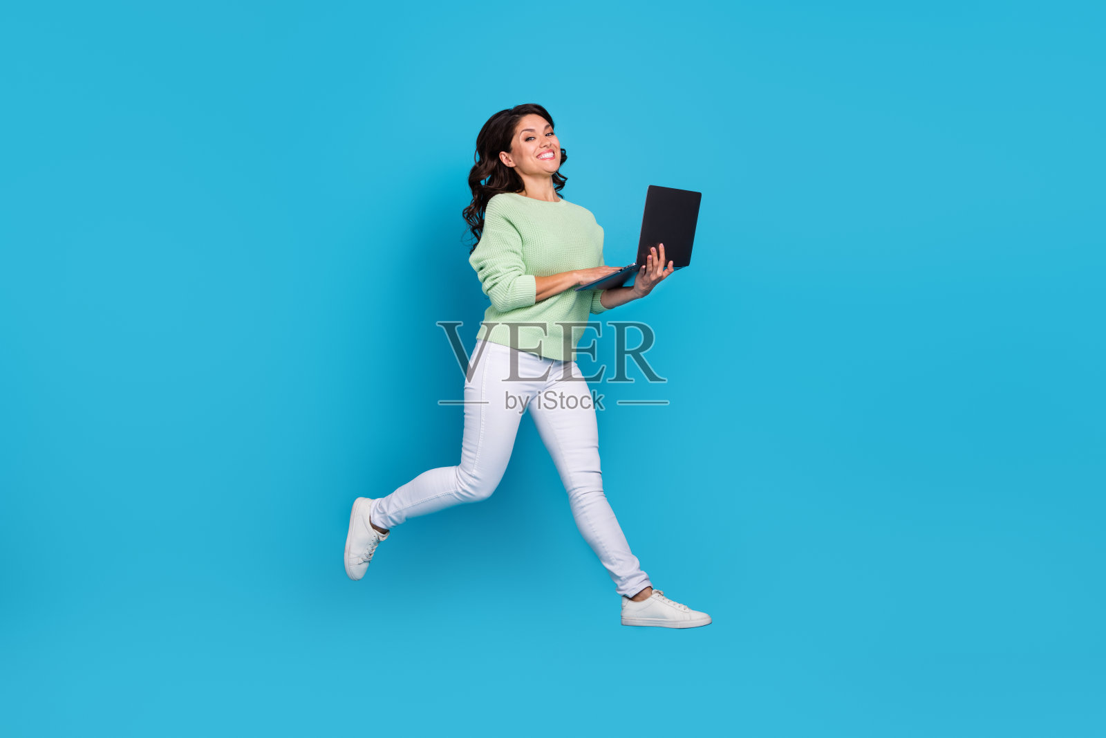 全身照，年轻的棕发女士穿着衬衫、裤子和运动鞋，跳跃着使用笔记本电脑，背景为蓝色。照片摄影图片