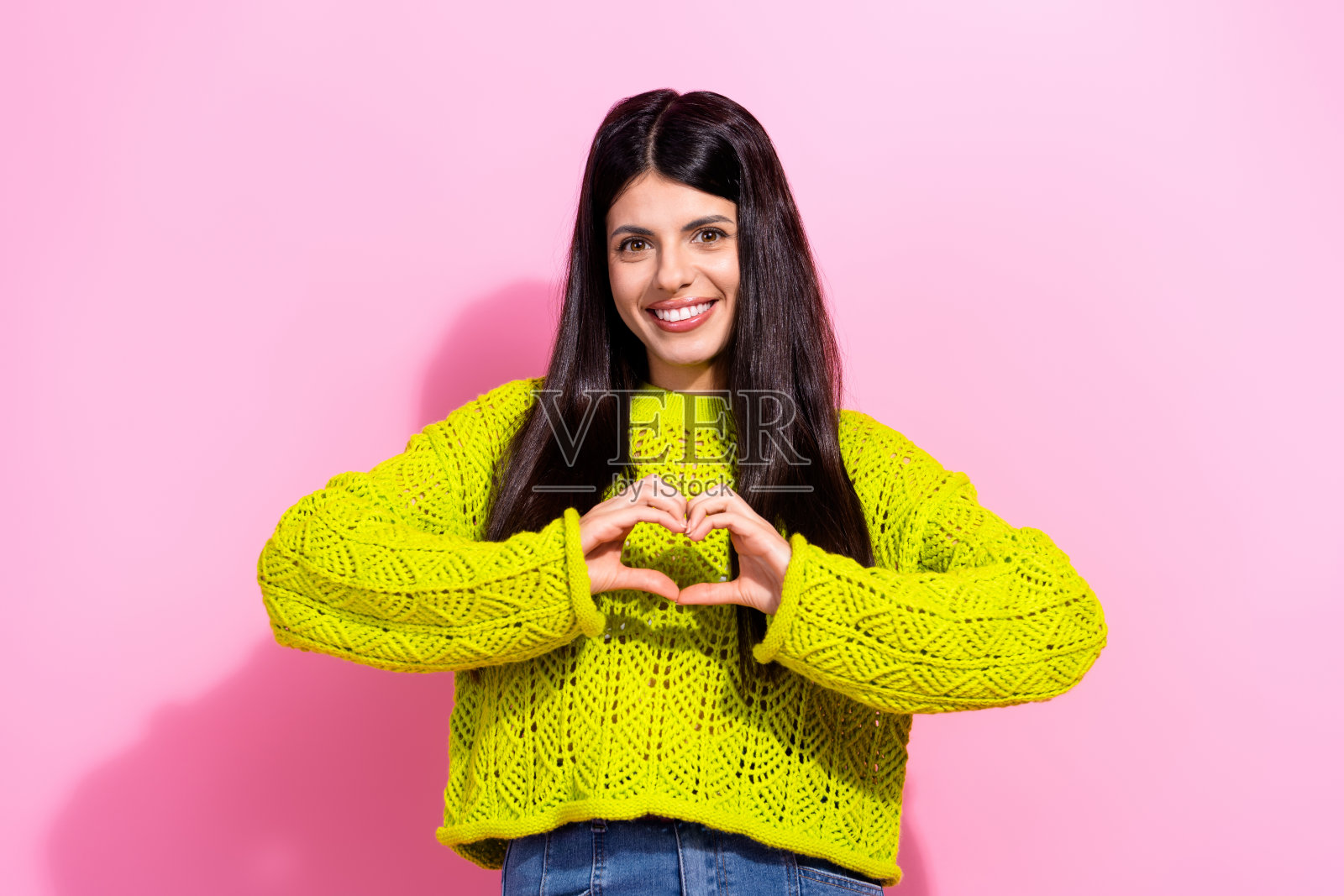 年轻女性穿着明亮的黄色毛衣，双手做出心形手势，面带微笑，背景是粉色，展现出魅力和快乐。照片摄影图片