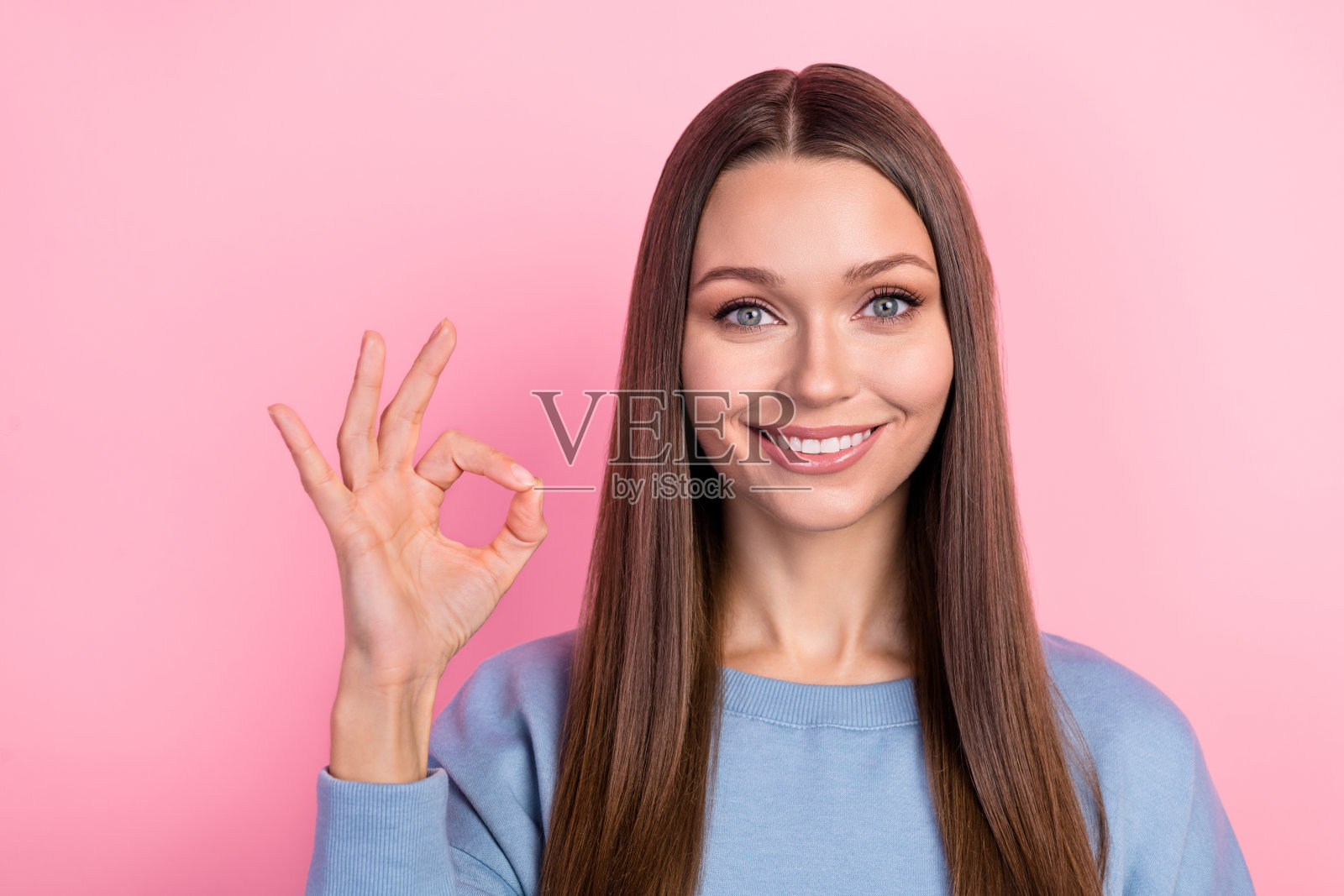 迷人的快乐棕发女孩在粉色柔和背景上做出“好”的手势。照片摄影图片