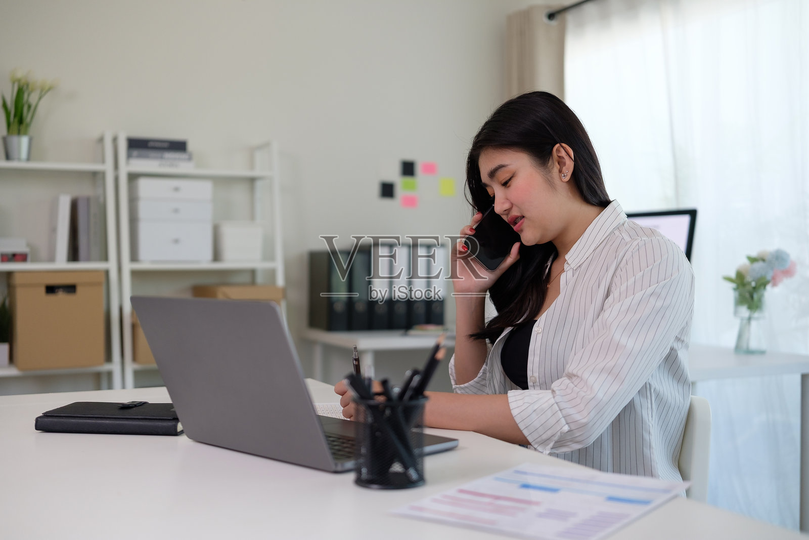 微笑的年轻女商人一边在桌子上查看财务文件，一边用手机通话。照片摄影图片