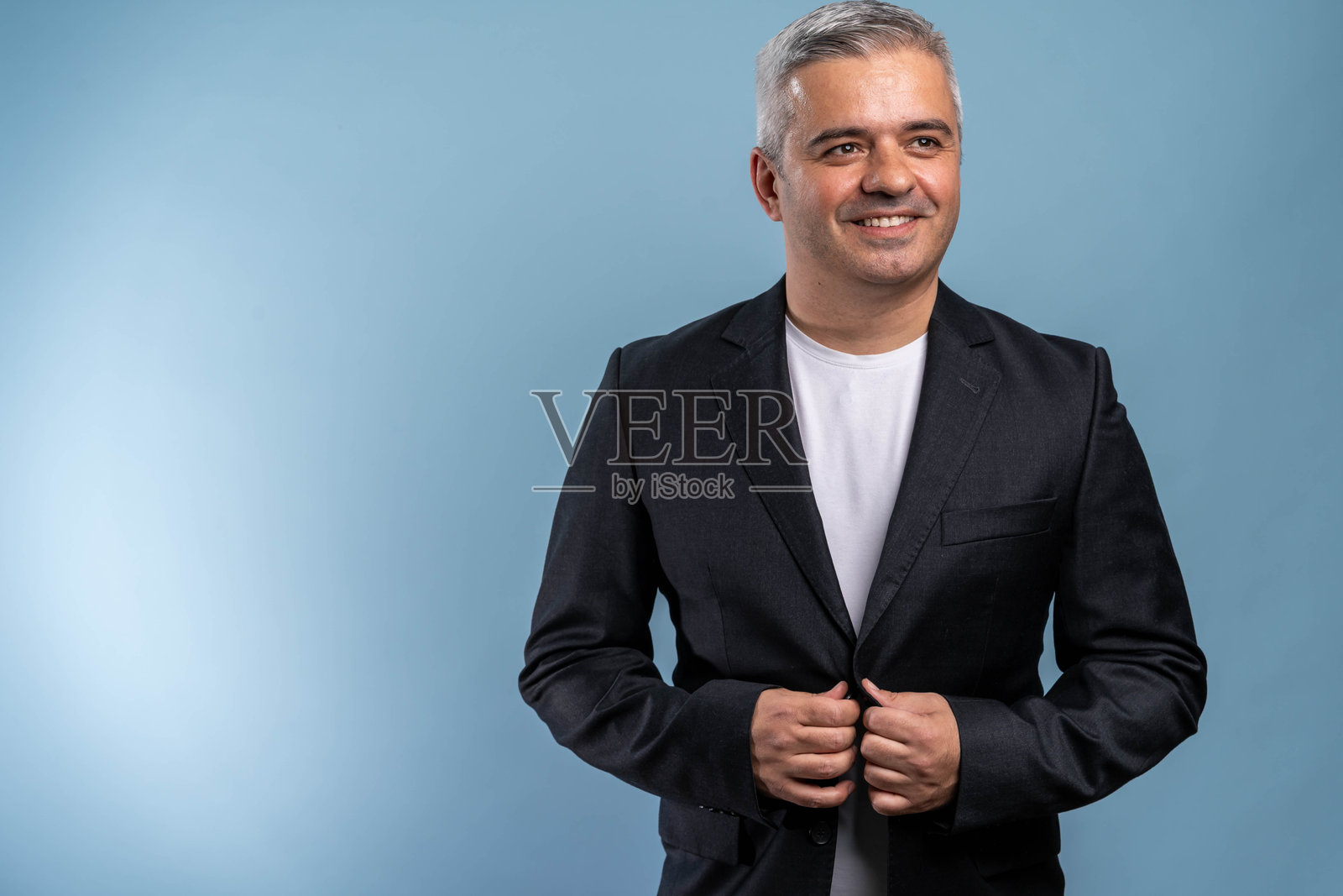 微笑的优雅中年男性经理穿着蓝色西装，自信地双手交叉，独自站在蓝色背景前。照片摄影图片
