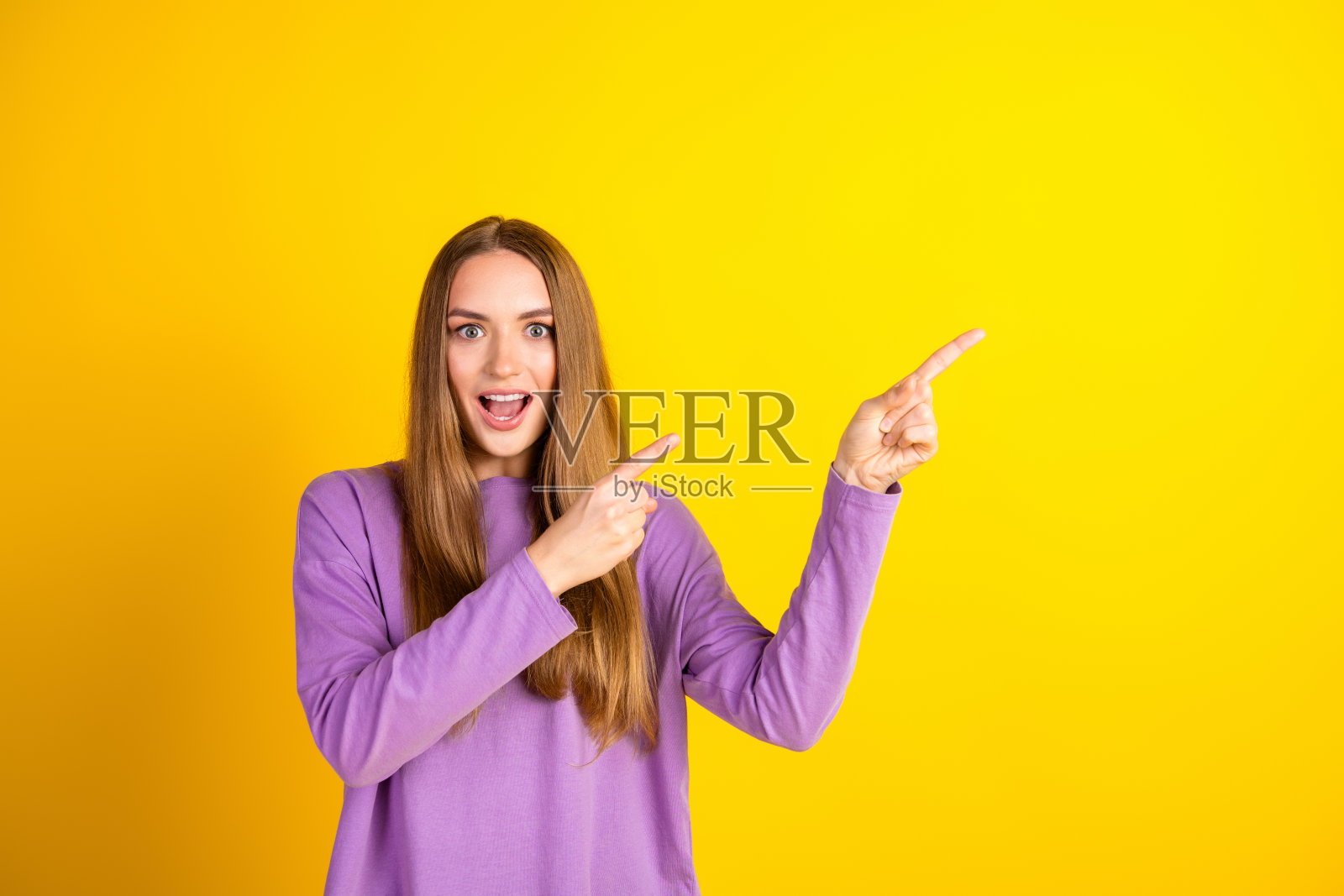 兴奋的年轻女性双手指向右侧，背景为鲜艳的黄色，展现出惊讶和快乐。照片摄影图片
