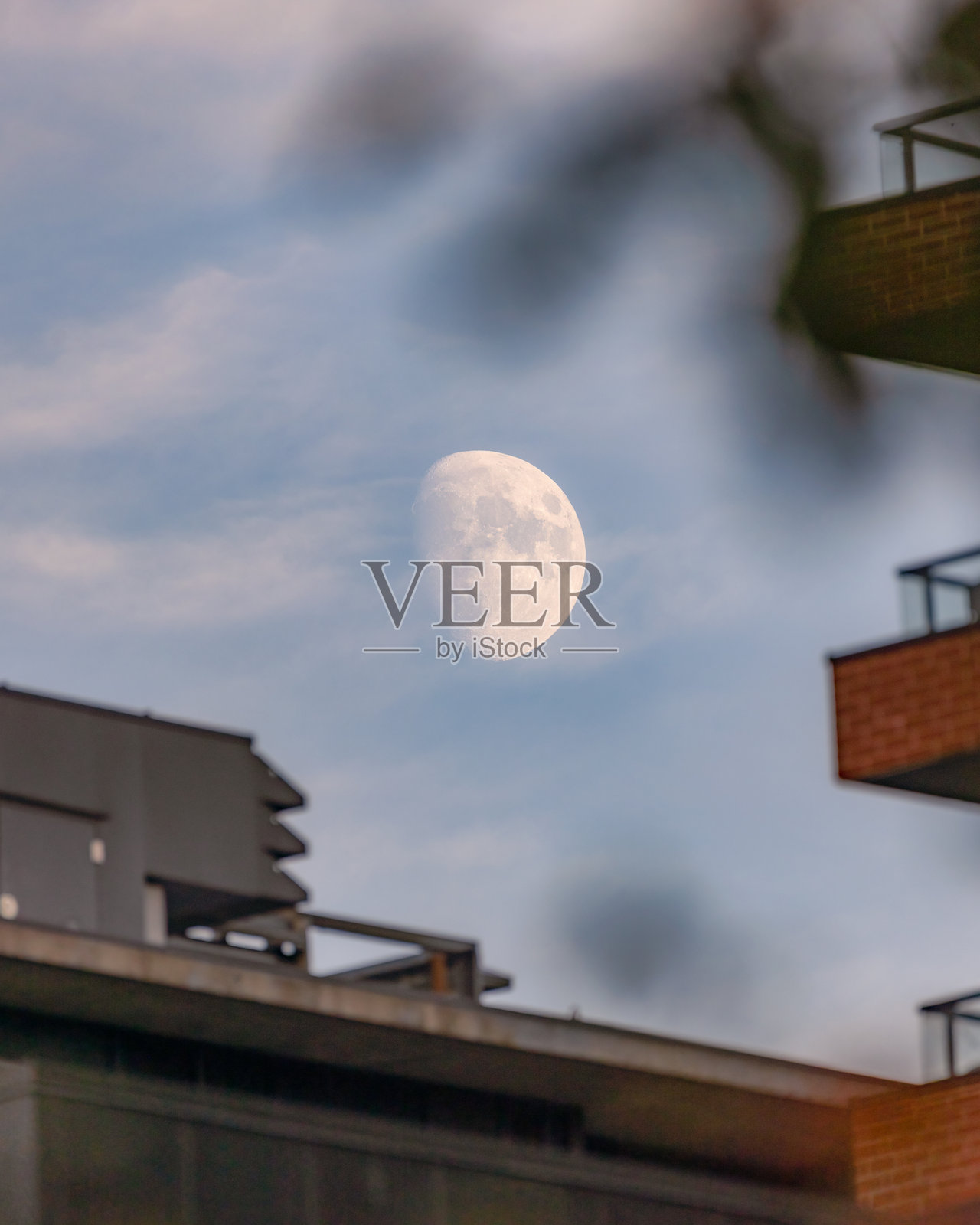 在朦胧的蓝天中，近满月的月亮升起在公寓楼上方。照片摄影图片