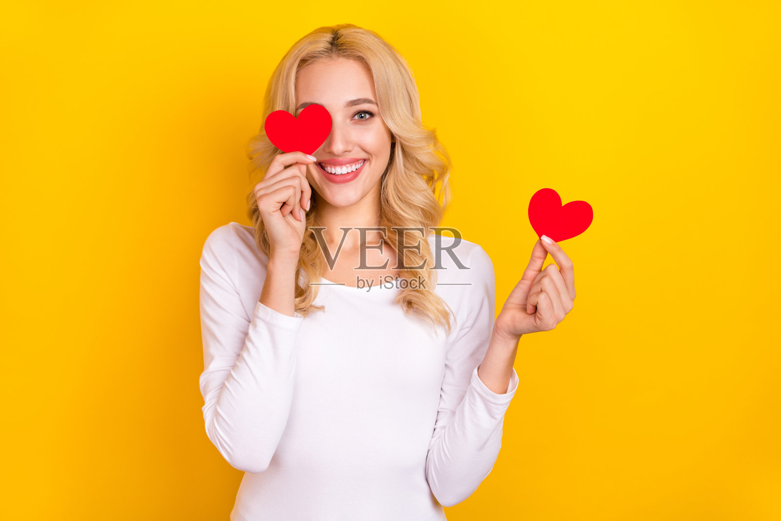 可爱的年轻女性穿着白衬衫，手捧小心形物品遮住眼睛，背景为黄色。照片摄影图片