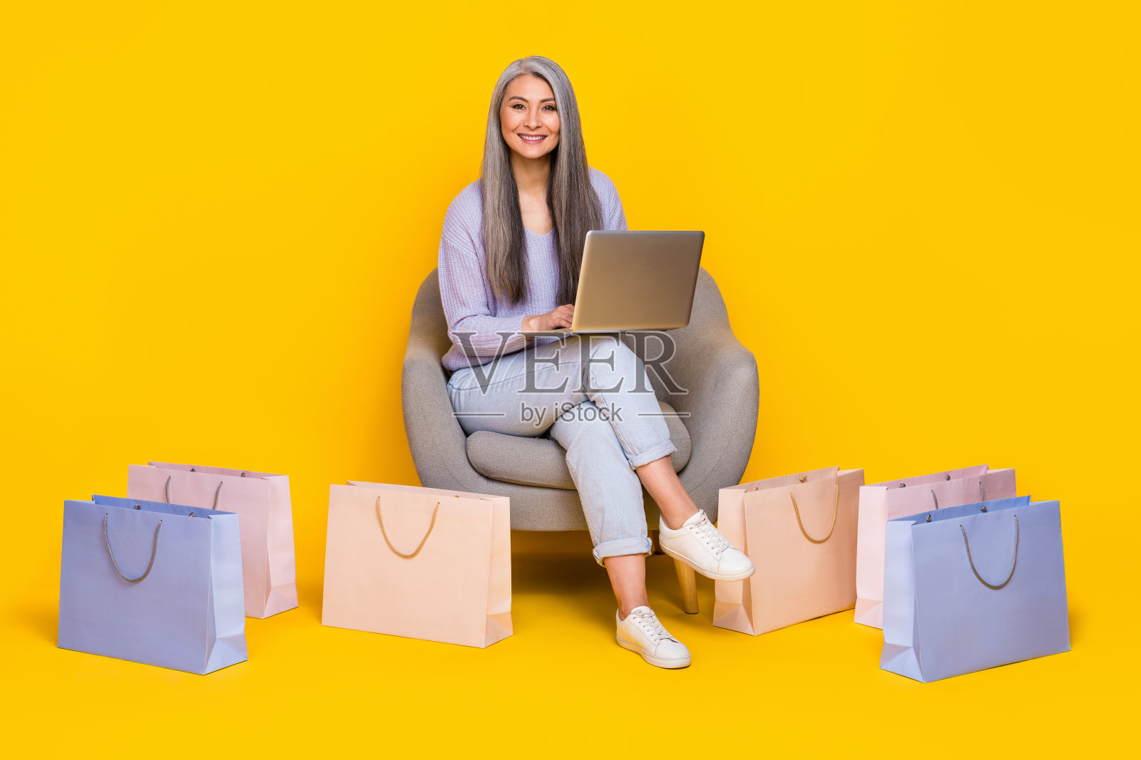 穿着毛衣的可爱退休女性坐在扶手椅上，使用现代设备打字，背景为黄色。照片摄影图片