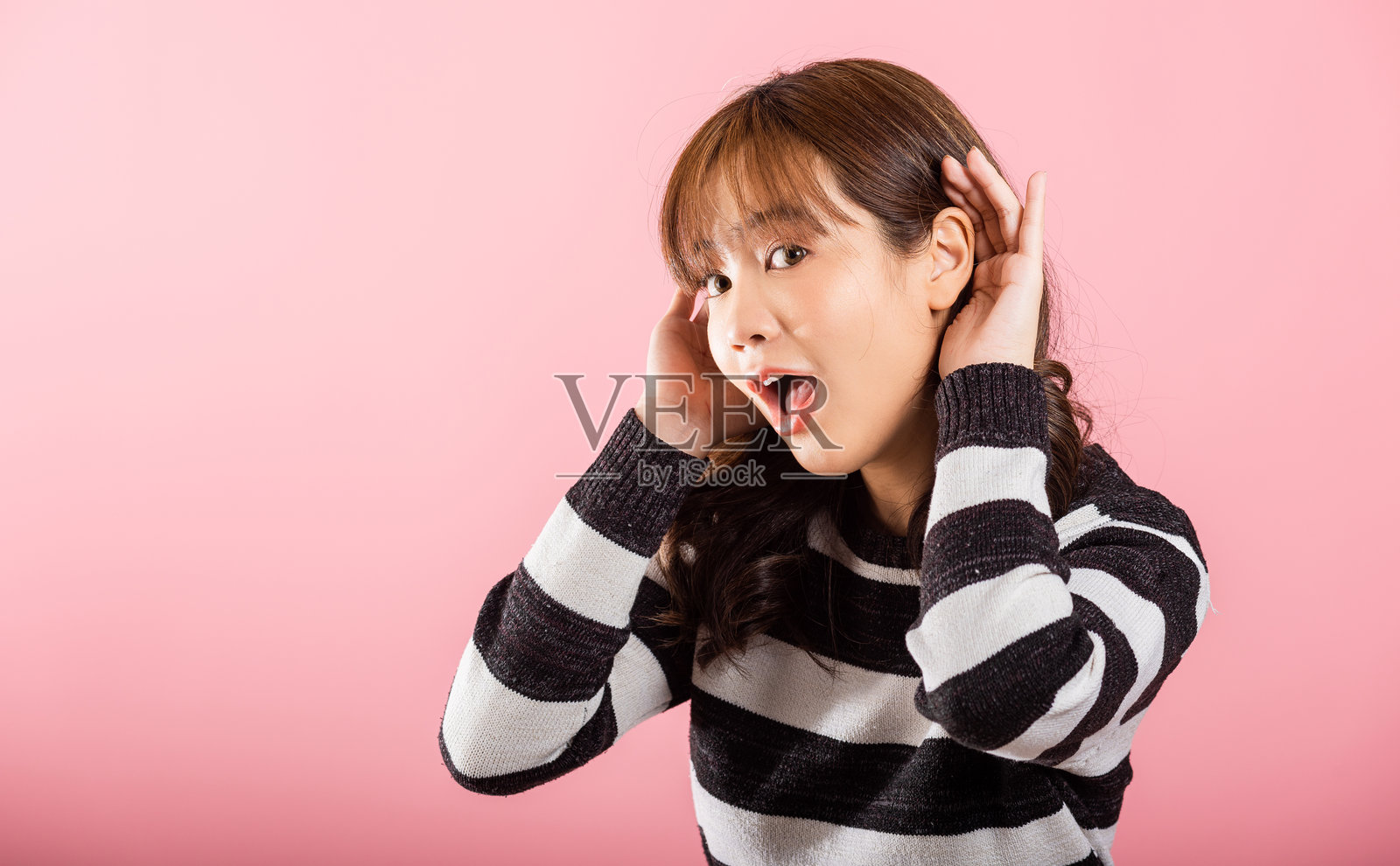 亚洲肖像，漂亮可爱的年轻女性，微笑着专注地倾听八卦，手放在耳边，工作室拍摄，粉色背景，留有空白空间。照片摄影图片