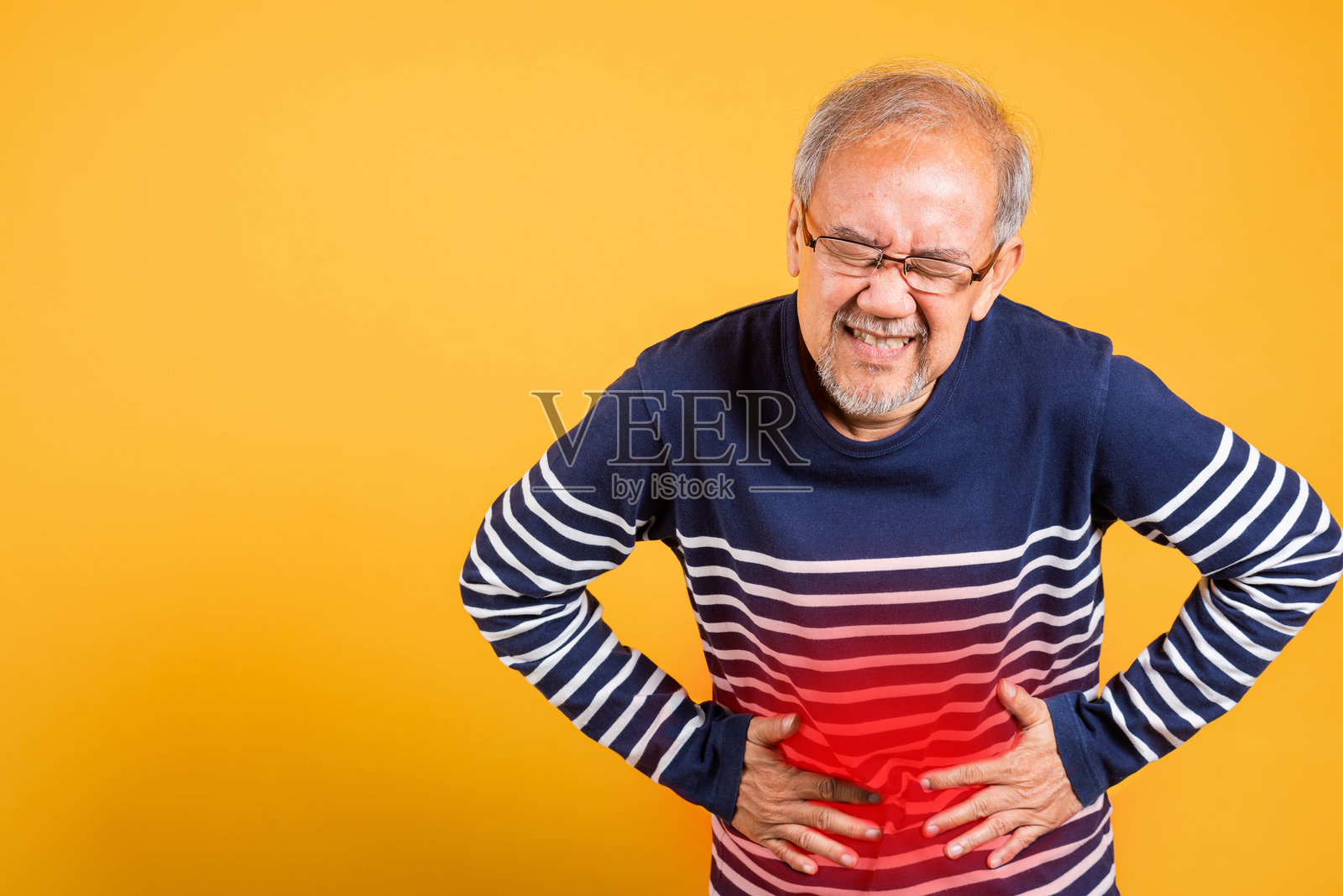 戴眼镜的亚洲老年男子双手放在腹部，感到胃痛，工作室拍摄，背景为黄色，老年人退休者感到腹部疼痛。照片摄影图片