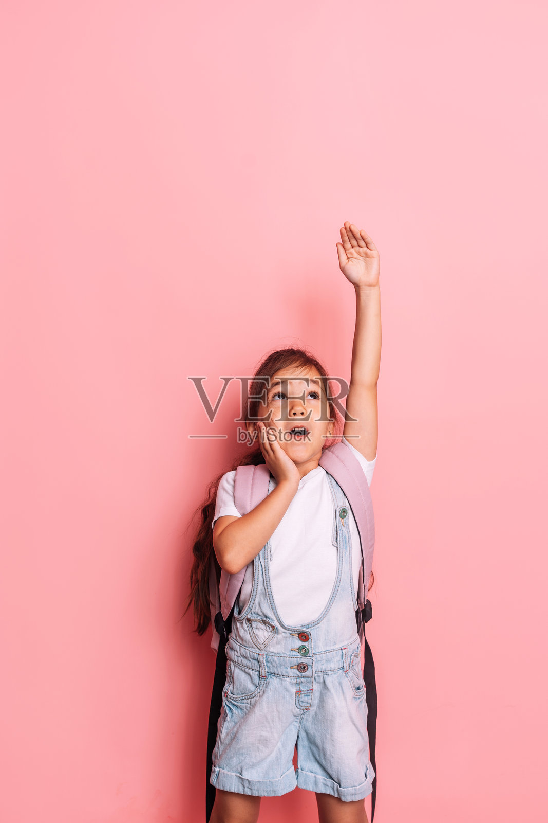 一个背着背包的女孩在粉色背景前举起了手。照片摄影图片