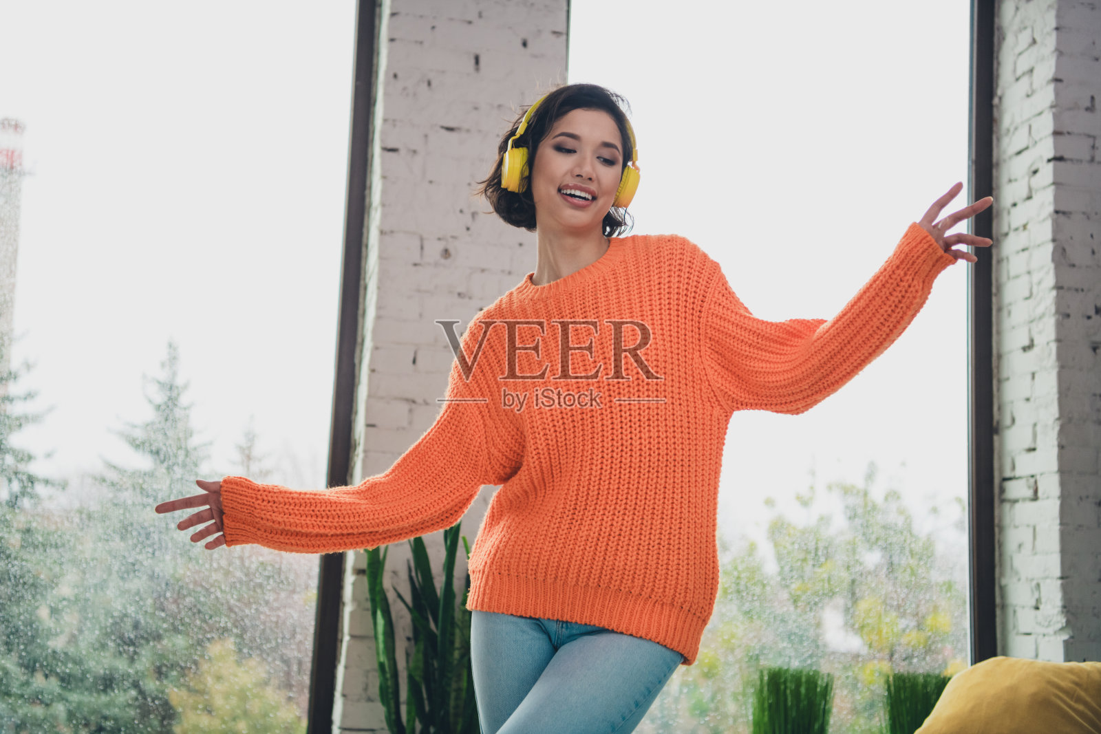 年轻女子穿着橙色毛衣，戴着耳机在家中享受音乐，伴随着明亮舒适的日子跳舞。照片摄影图片