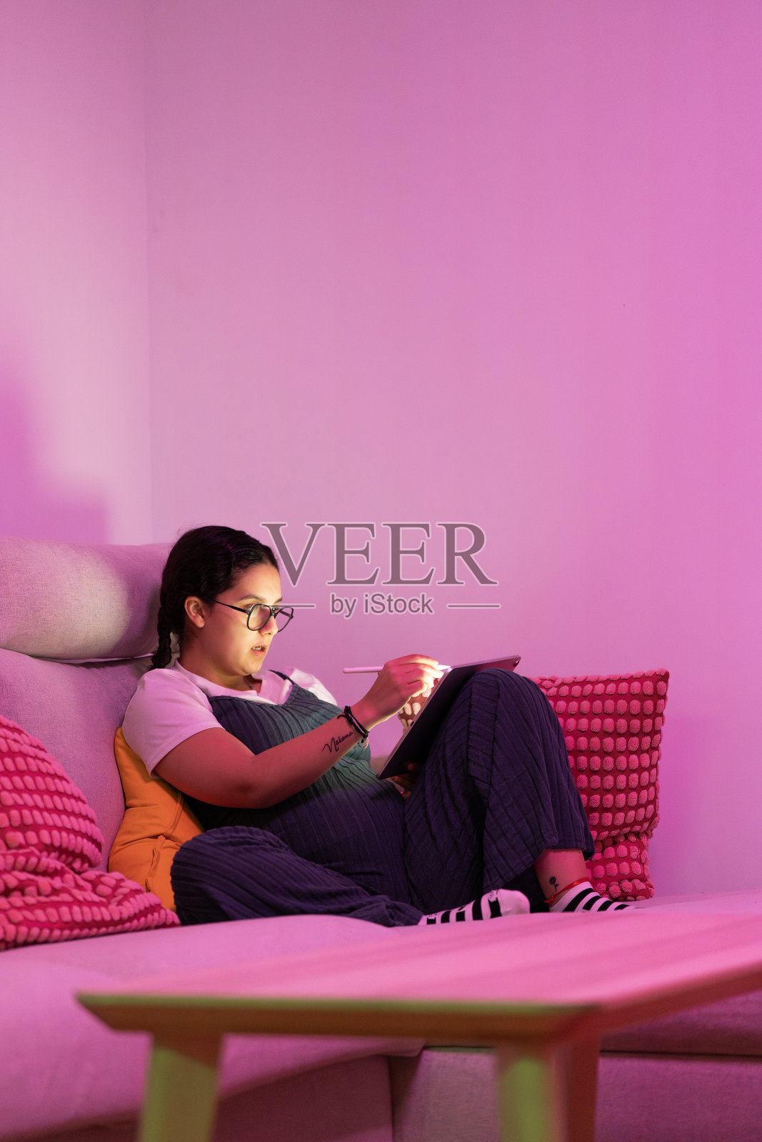 怀孕的女人在粉色灯光的房间里放松地坐在沙发上，用平板电脑绘画照片摄影图片