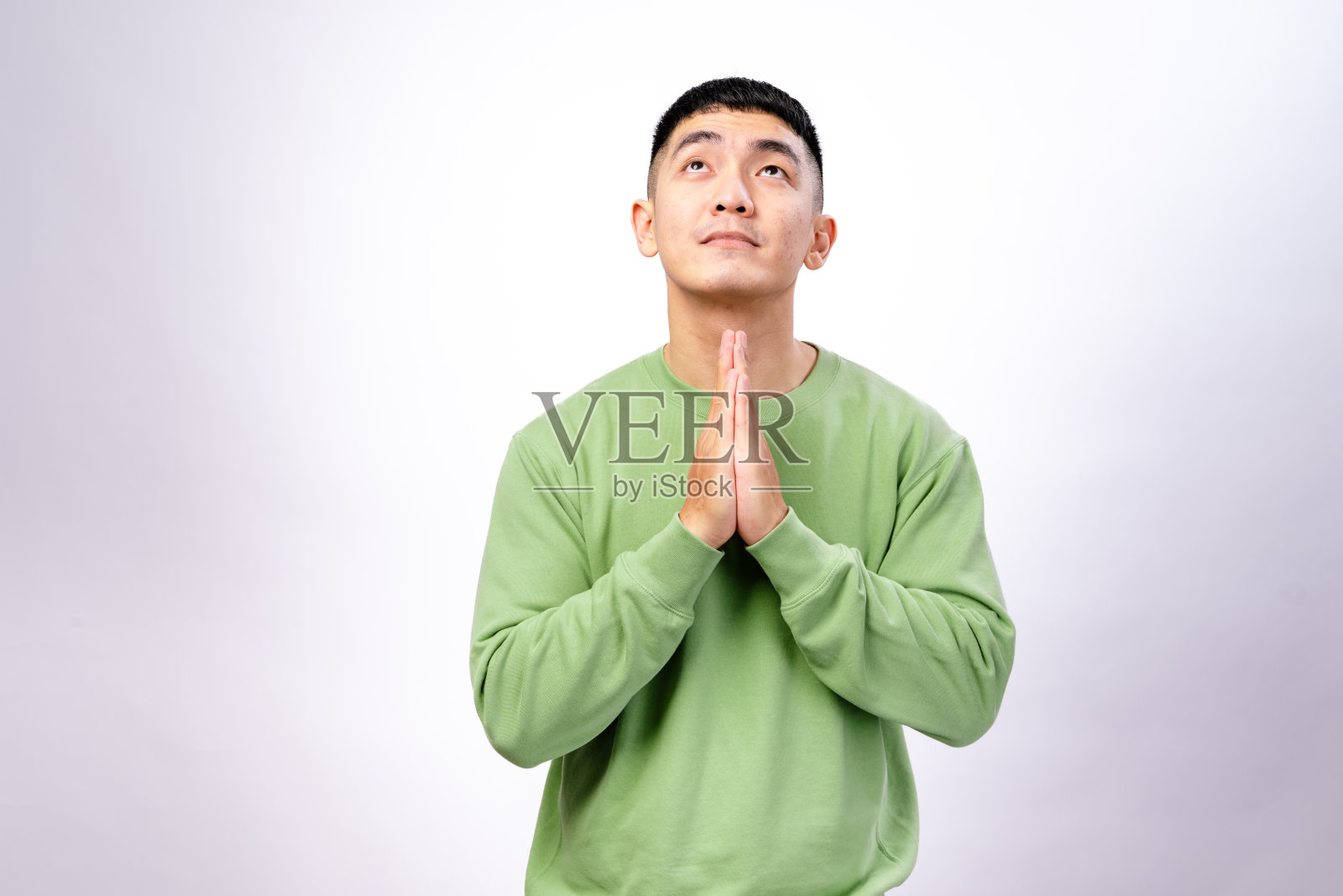 一位穿着绿色卫衣的亚洲男子站在白色背景前，双手合十，表达感激、希望或冥想的姿态。照片摄影图片