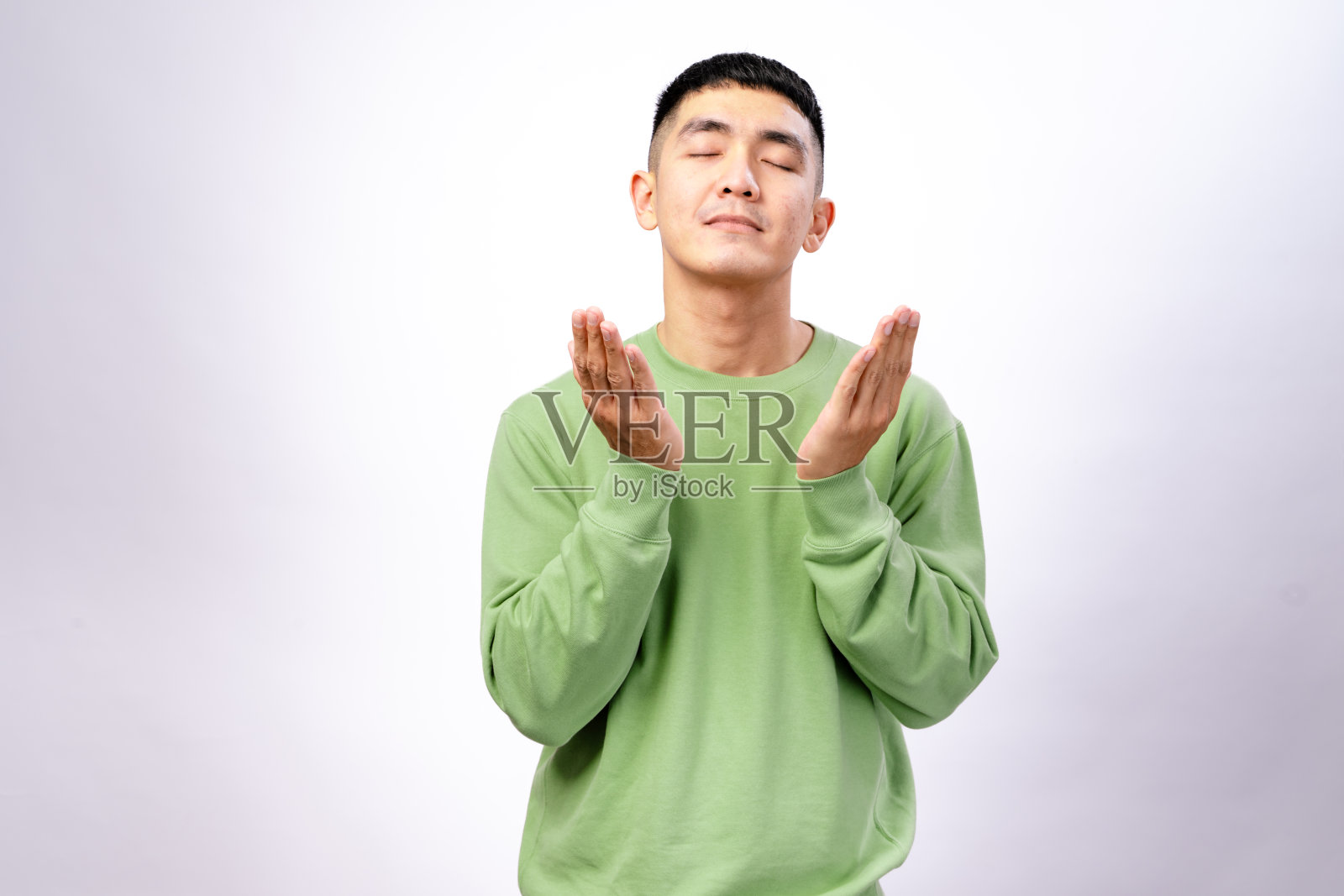 一位穿着绿色卫衣的亚洲男子站在白色背景前，仰望天空，双手举起，做出祈祷或充满希望的姿势，表达感激、信仰或沉思。照片摄影图片