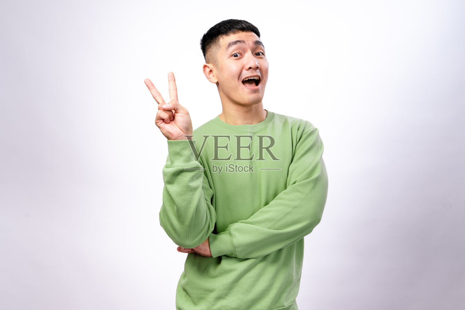 一位穿着绿色卫衣的亚洲男子用右手做出和平手势，微微一笑，双臂交叉，站在白色背景前，展现出自信的姿态。照片摄影图片