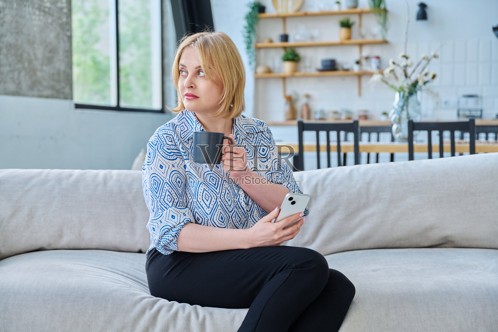 中年金发女性坐在客厅沙发上，手持智能手机和杯子。照片摄影图片