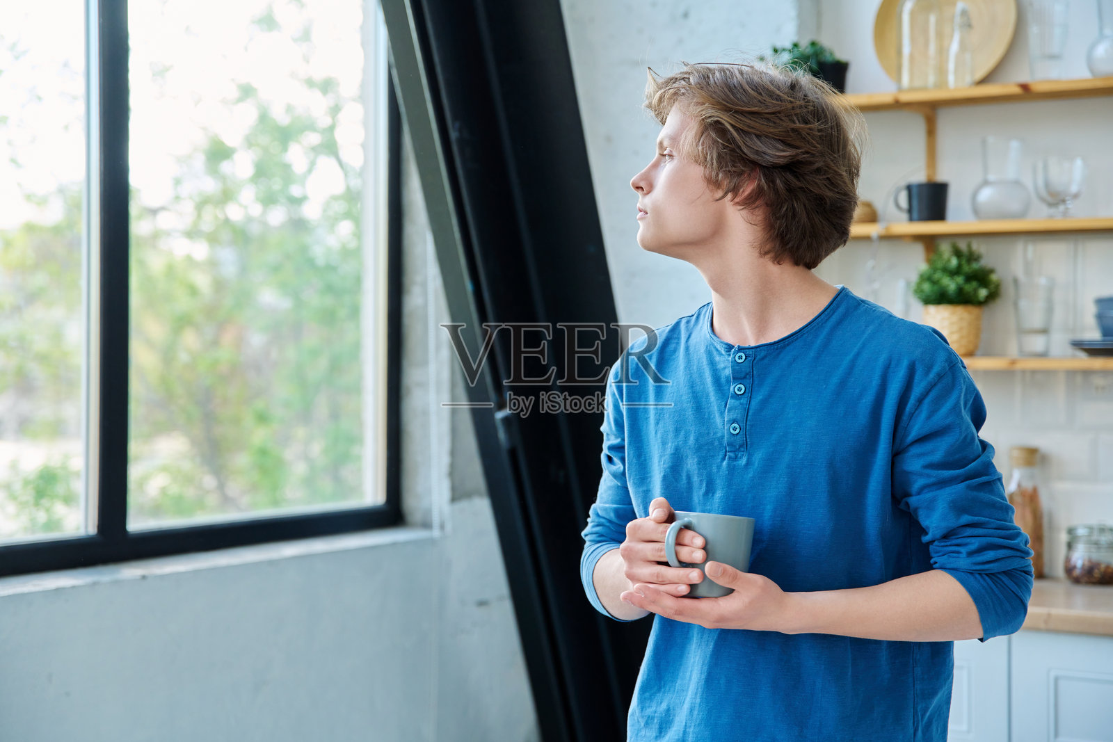 手握杯子的严肃少年在家居环境中，望向窗外照片摄影图片