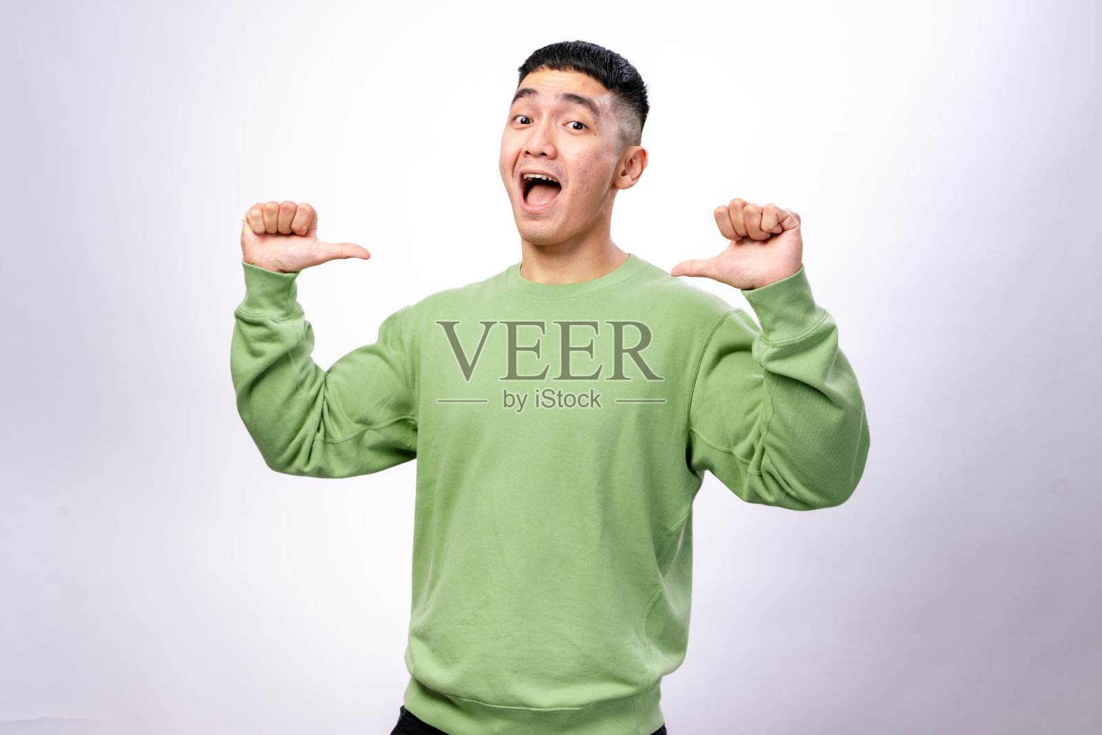 一位穿着绿色卫衣的亚洲男子站在白色背景前，自信地微笑，同时用双拇指指向自己，表现出骄傲和自信的姿态。照片摄影图片