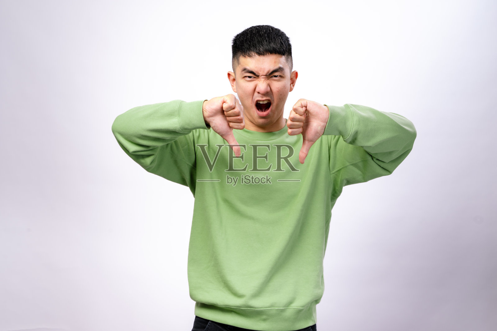一位穿着绿色卫衣的亚洲男子站在白色背景前，做出双手竖大拇指向下的手势，面露强烈愤怒的表情，表现出强烈的不满或沮丧。照片摄影图片