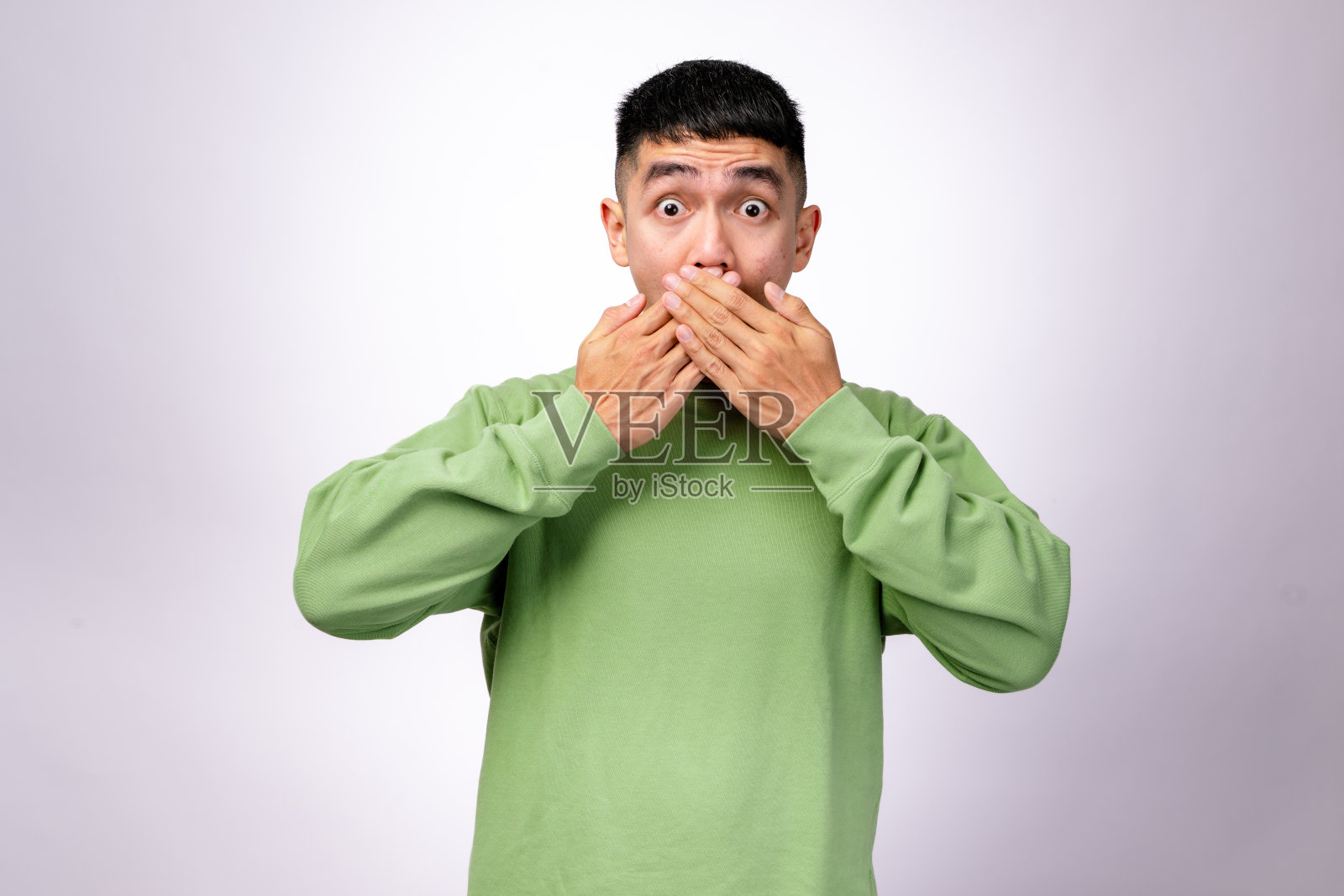 一位穿着绿色卫衣的亚洲男子用双手捂住嘴，眼睛睁得大大的，流露出惊讶或震惊的表情。他的表情传达出秘密和领悟，背景为纯白色。照片摄影图片
